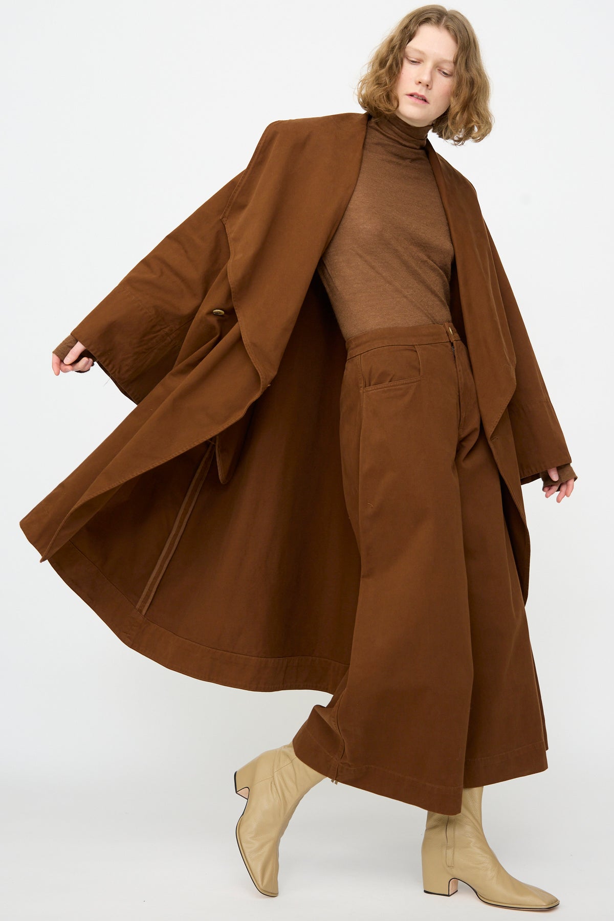A person wearing the Lauren Manoogian Planes Coat in Rust—a mid-length, oversized cotton coat—styled with a brown turtleneck, wide-leg brown pants, and beige heeled boots poses against a plain white background.