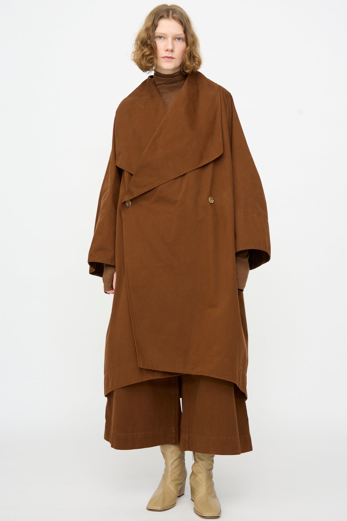 A person stands against a plain background, wearing the Lauren Manoogian Planes Coat in Rust—a long, oversized mid-length cotton coat—paired with brown wide-leg pants, a brown turtleneck, and beige boots.