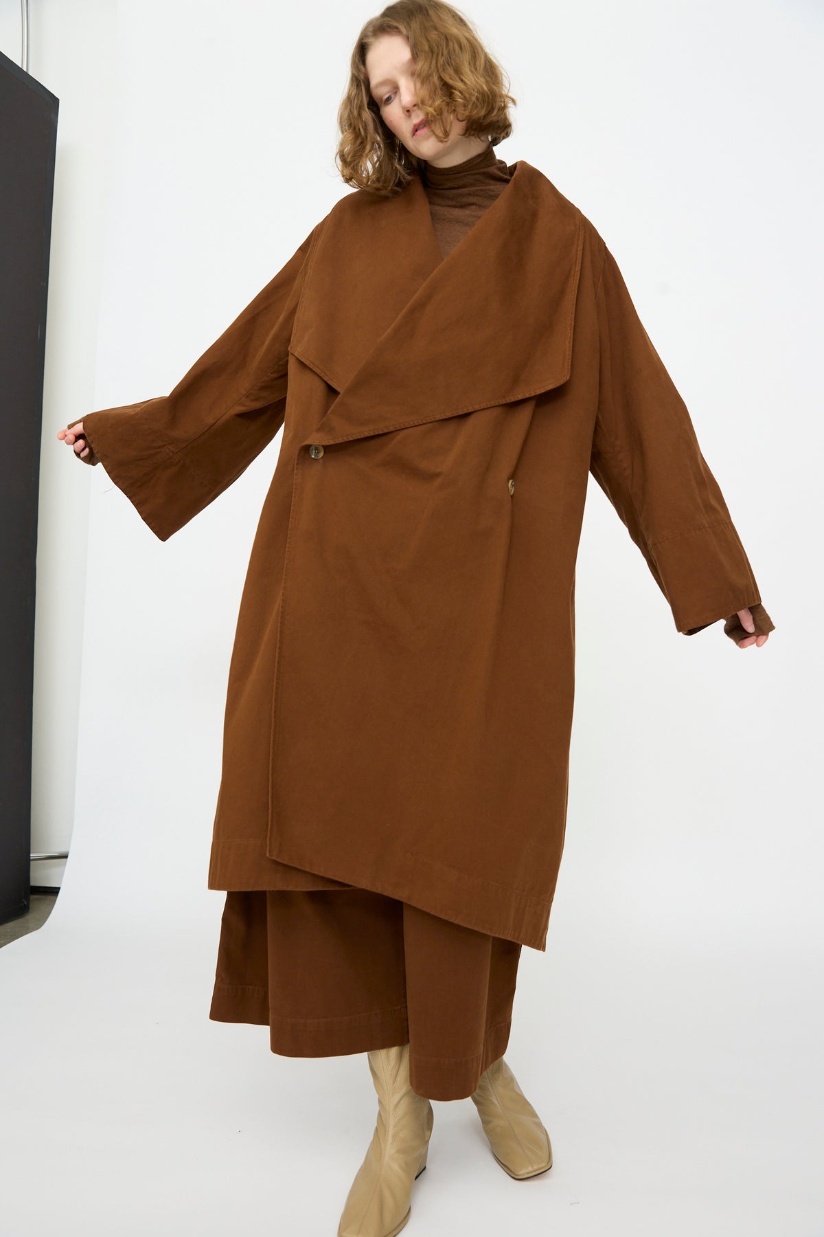 A person stands against a plain white background wearing the Lauren Manoogian Planes Coat in Rust, paired with a matching brown turtleneck, brown wide-leg pants, and beige boots.