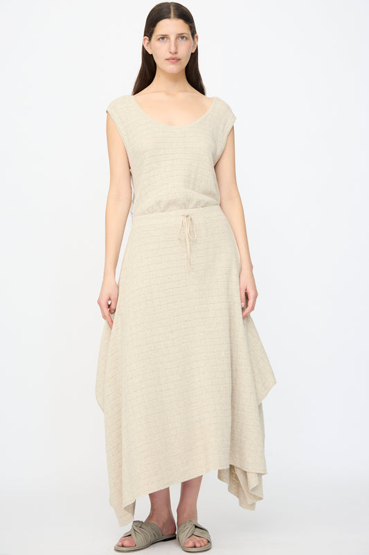 A woman faces forward against a plain white background, wearing the Lauren Manoogian Planes Skirt in Flax—sleeveless with a drawstring waist and handkerchief hem—paired with light-colored slide sandals.