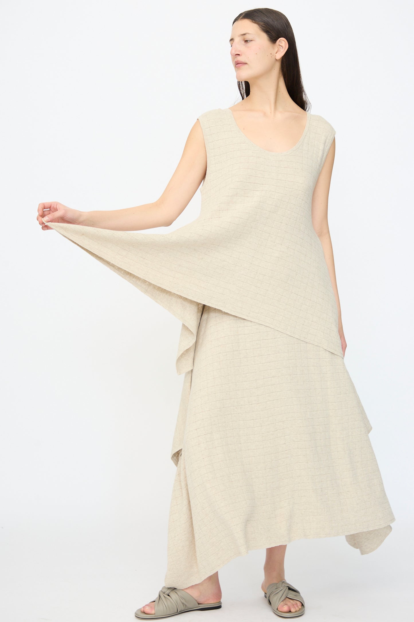 A woman models the Lauren Manoogian Planes Tank in Flax, a loose, textured beige dress. She stands against a plain white background, holding one side of the dress and pairing it with flat knotted sandals for an effortlessly chic look.