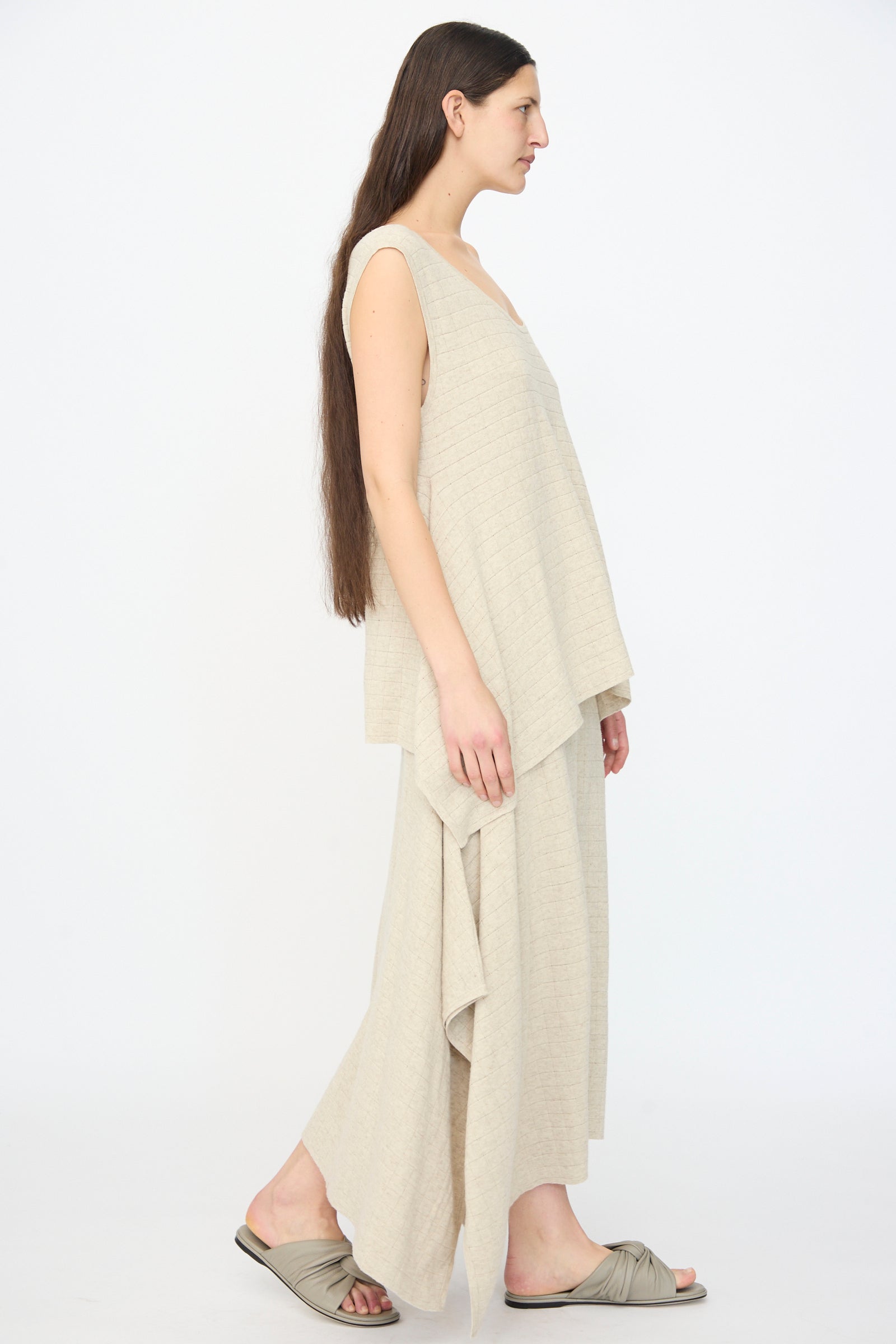 A woman in profile wears the Lauren Manoogian Planes Tank in Flax, a sleeveless, relaxed-fit beige linen top paired with a matching skirt and beige slide sandals, set against a plain white background.