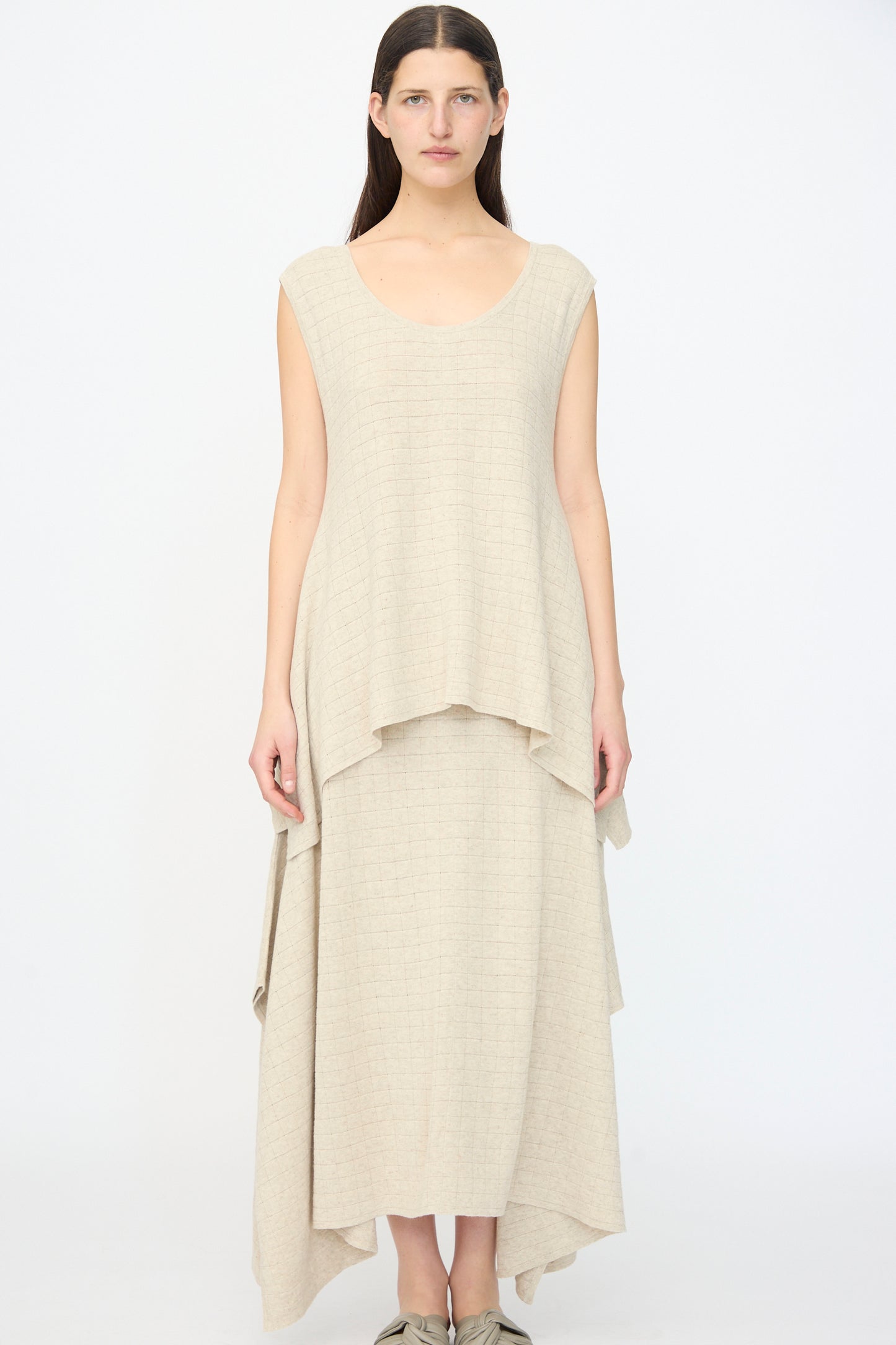 A woman stands facing forward against a white background, wearing the Lauren Manoogian Planes Tank in Flax—a relaxed fit, sleeveless, beige layered maxi dress with an uneven hem.