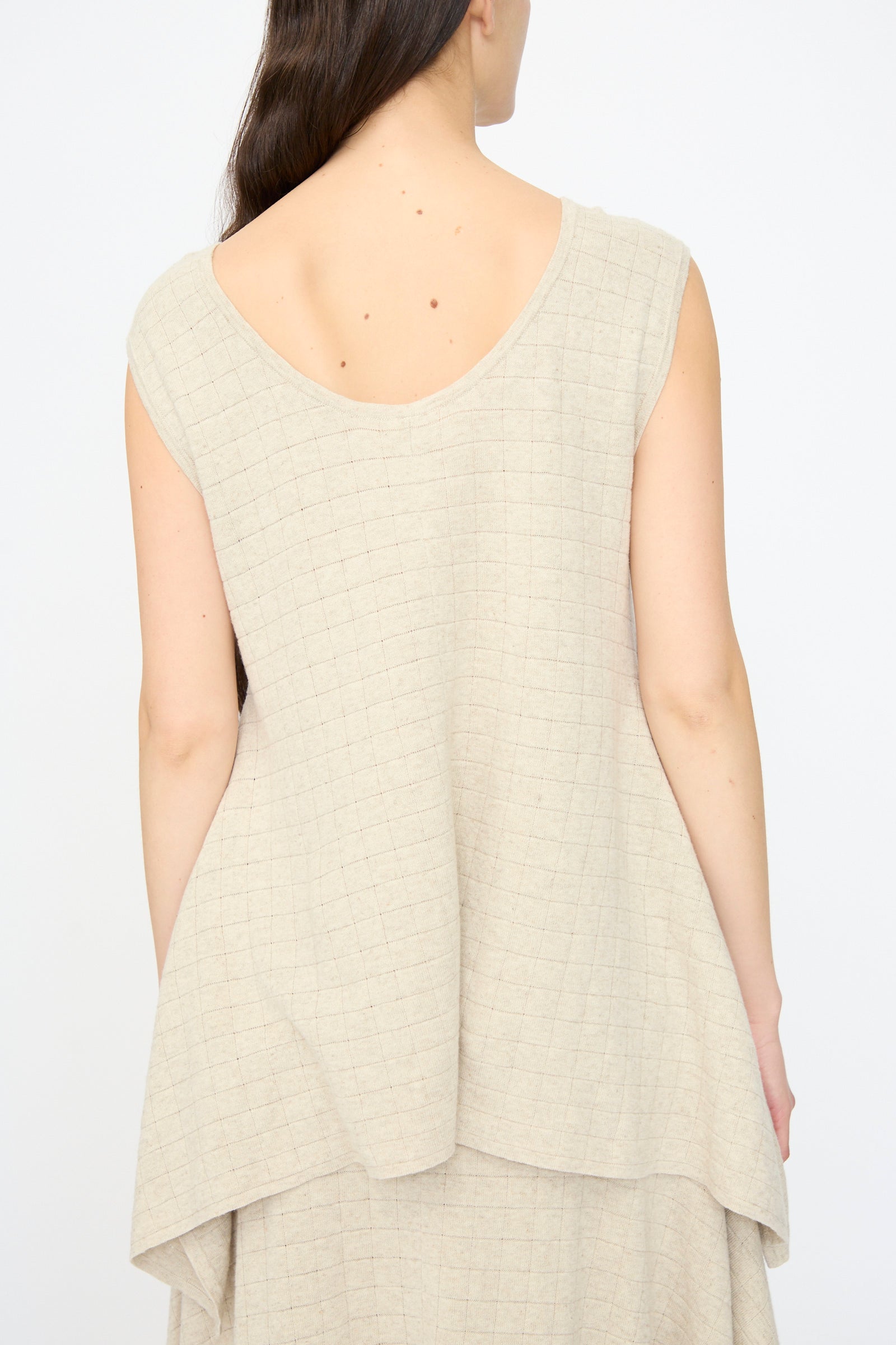 A woman with long brown hair is seen from the back wearing the Lauren Manoogian Planes Tank in Flax, a relaxed fit, sleeveless beige linen top featuring a grid pattern, set against a plain background.