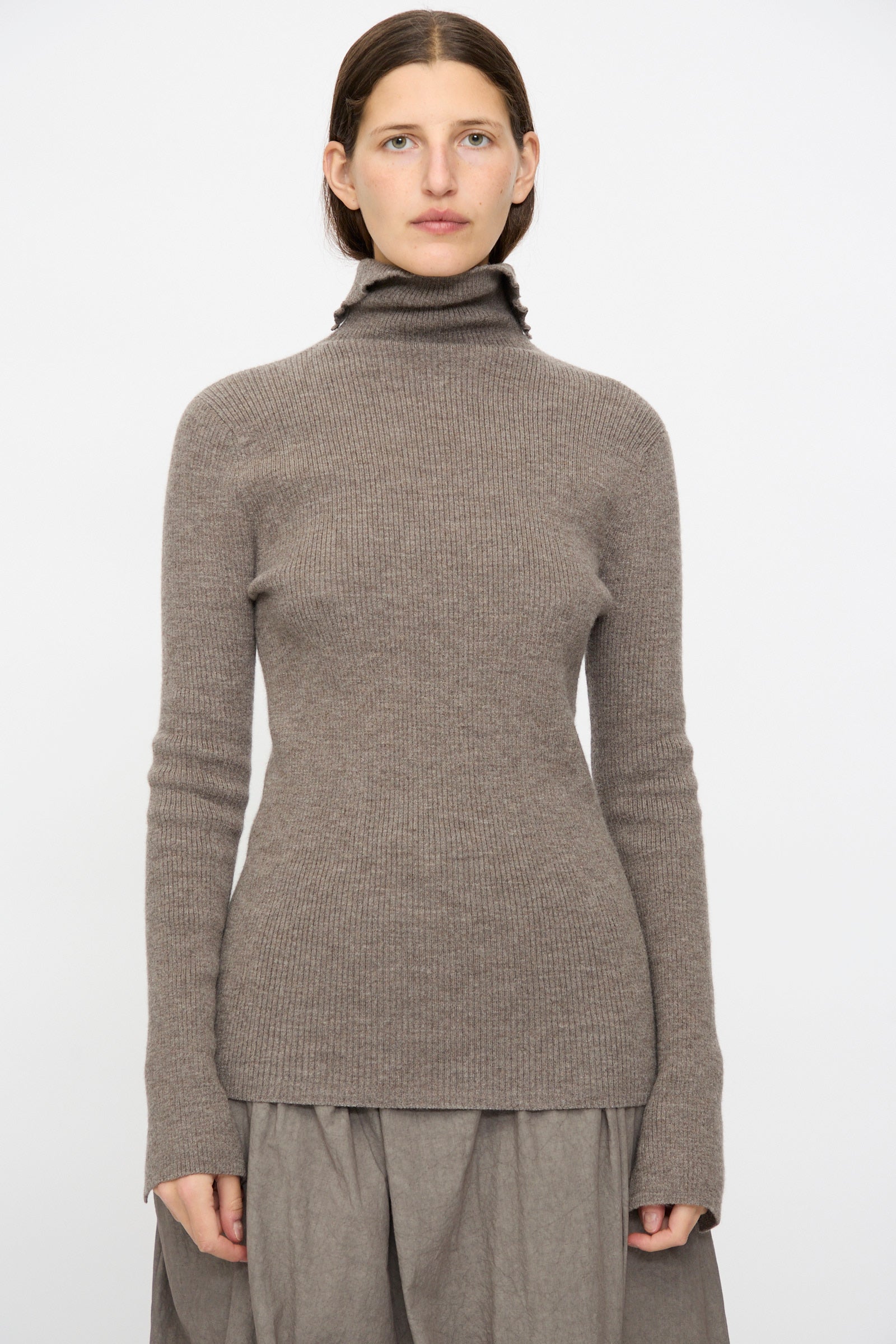 A person faces forward against a plain white background, wearing the Lauren Manoogian Rib Split Turtleneck in Cinder and a gray skirt.