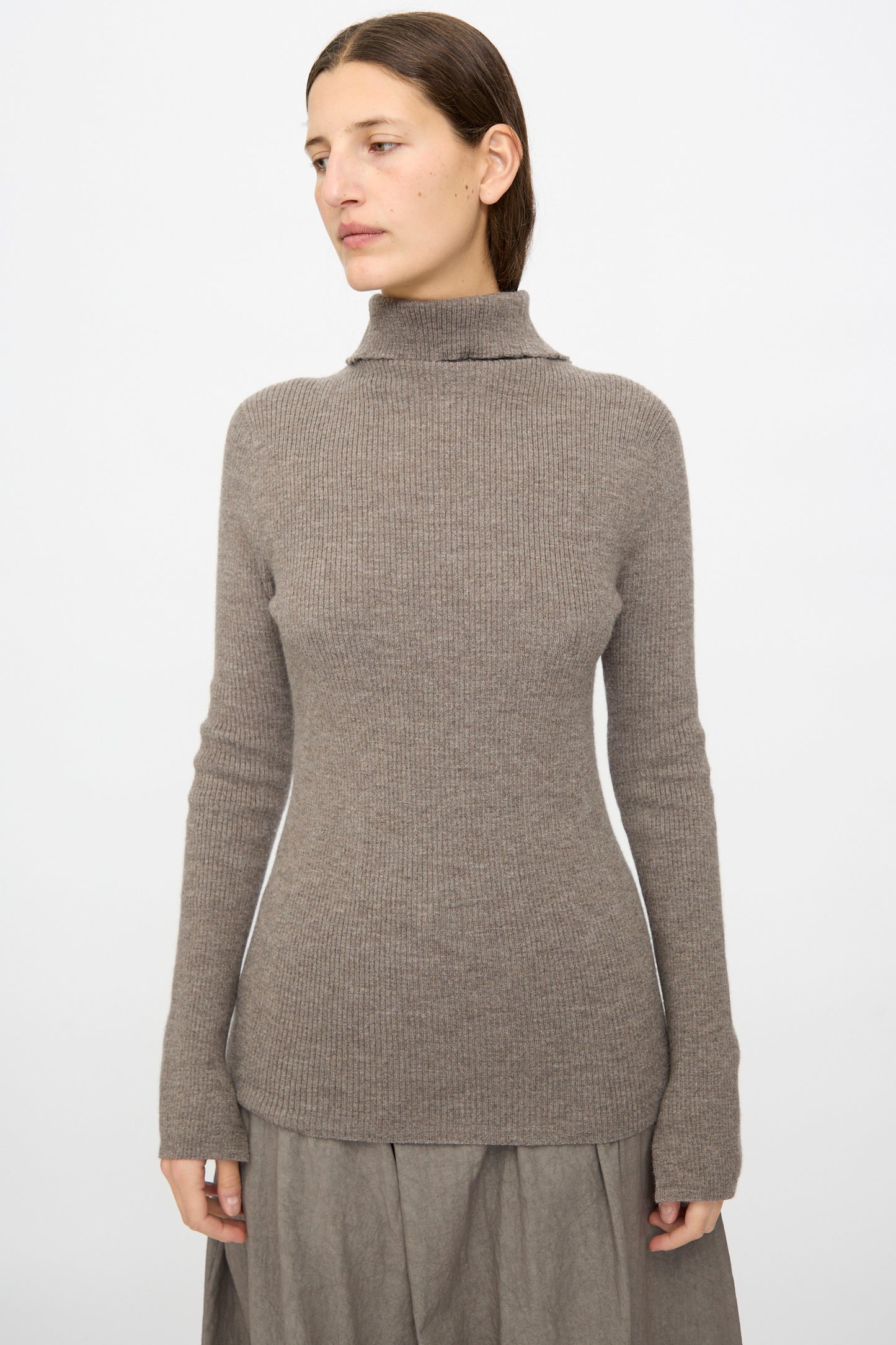 A woman wearing the Lauren Manoogian Rib Split Turtleneck in Cinder stands against a plain white background, paired with a gray skirt and facing slightly to the side.