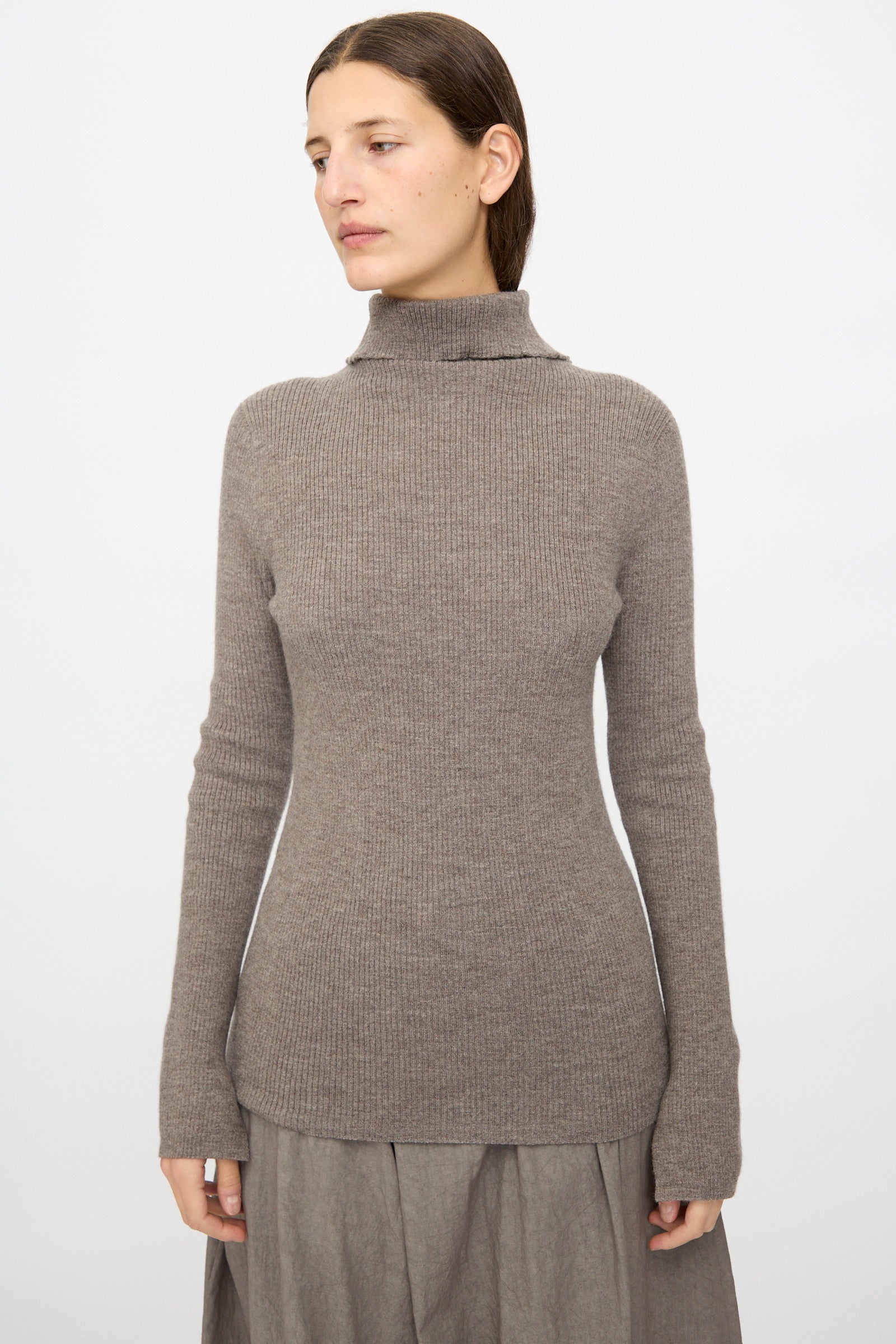 A woman wearing the Lauren Manoogian Rib Split Turtleneck in Cinder stands against a plain white background, paired with a gray skirt and facing slightly to the side.