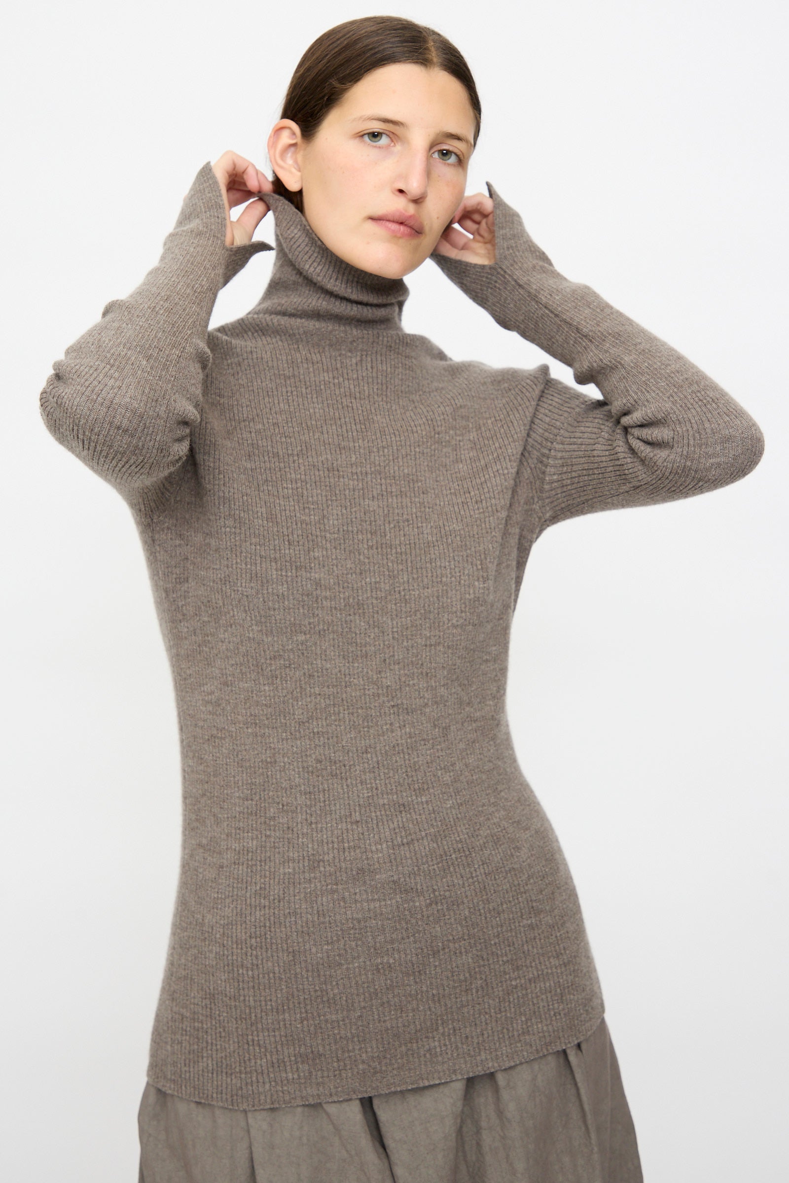A person wearing the Lauren Manoogian Rib Split Turtleneck in Cinder and a grey skirt adjusts the collar, standing against a plain light background.