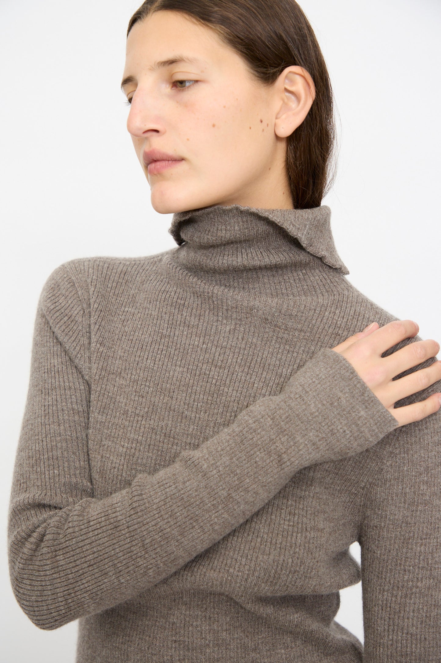Person with straight brown hair models the Lauren Manoogian Rib Split Turtleneck in Cinder, looking to the side with one hand on their shoulder, set against a plain background.