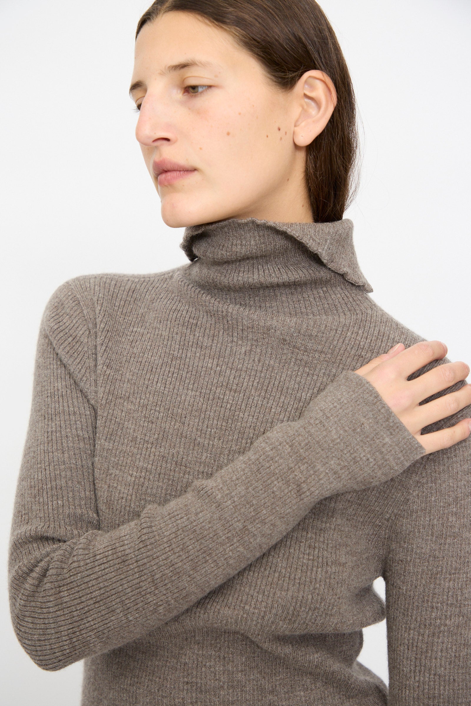 Person with straight brown hair models the Lauren Manoogian Rib Split Turtleneck in Cinder, looking to the side with one hand on their shoulder, set against a plain background.