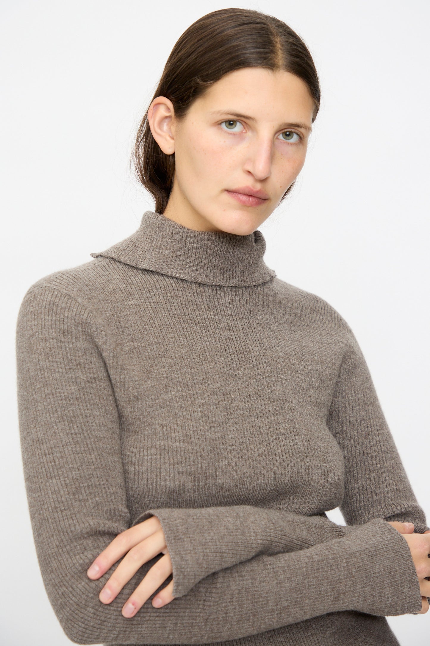 A person with straight brown hair wears the Lauren Manoogian Rib Split Turtleneck in Cinder, featuring soft ribbed merino wool, standing with arms crossed and facing the camera against a plain background.