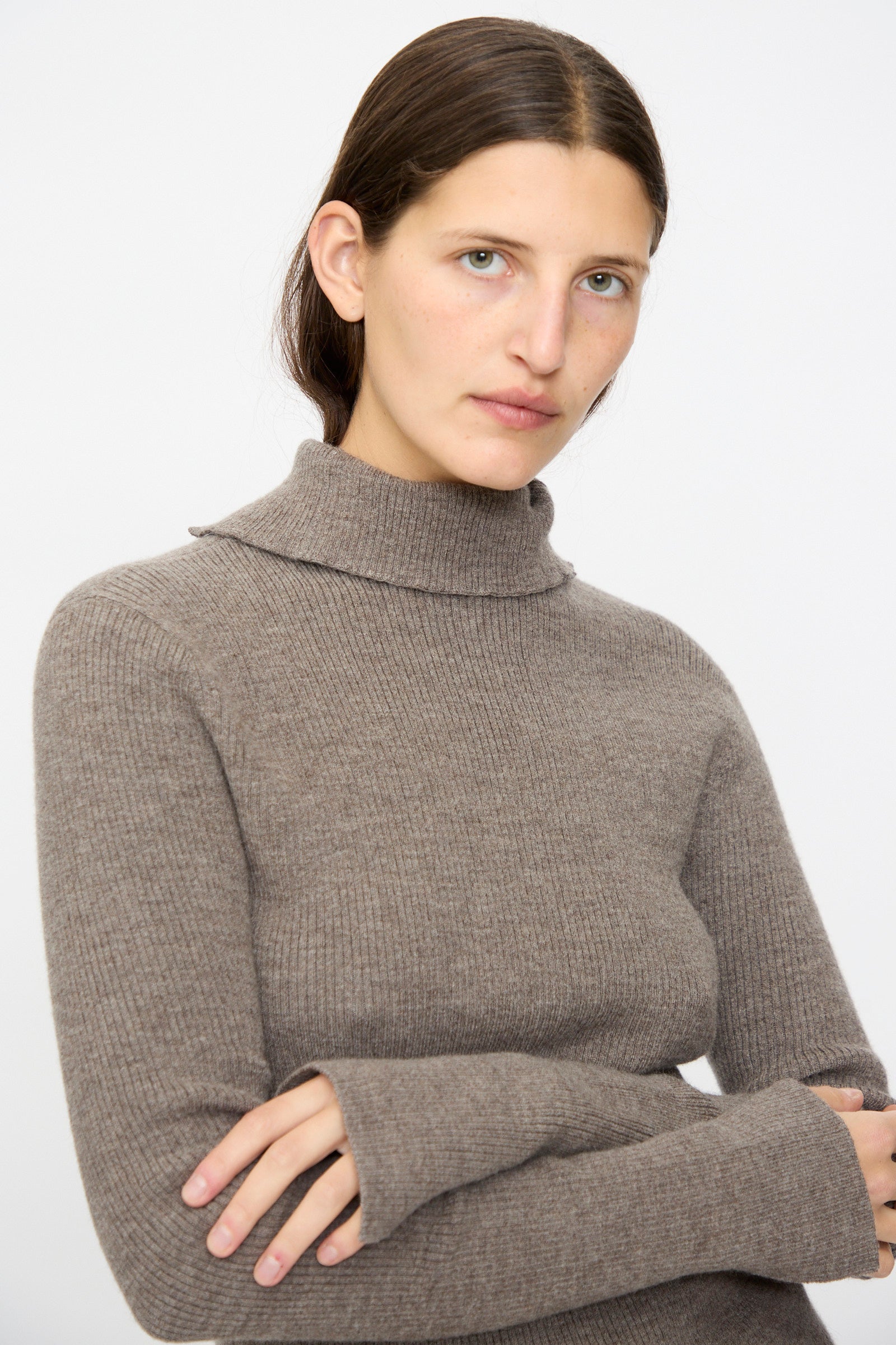 A person with straight brown hair wears the Lauren Manoogian Rib Split Turtleneck in Cinder, featuring soft ribbed merino wool, standing with arms crossed and facing the camera against a plain background.
