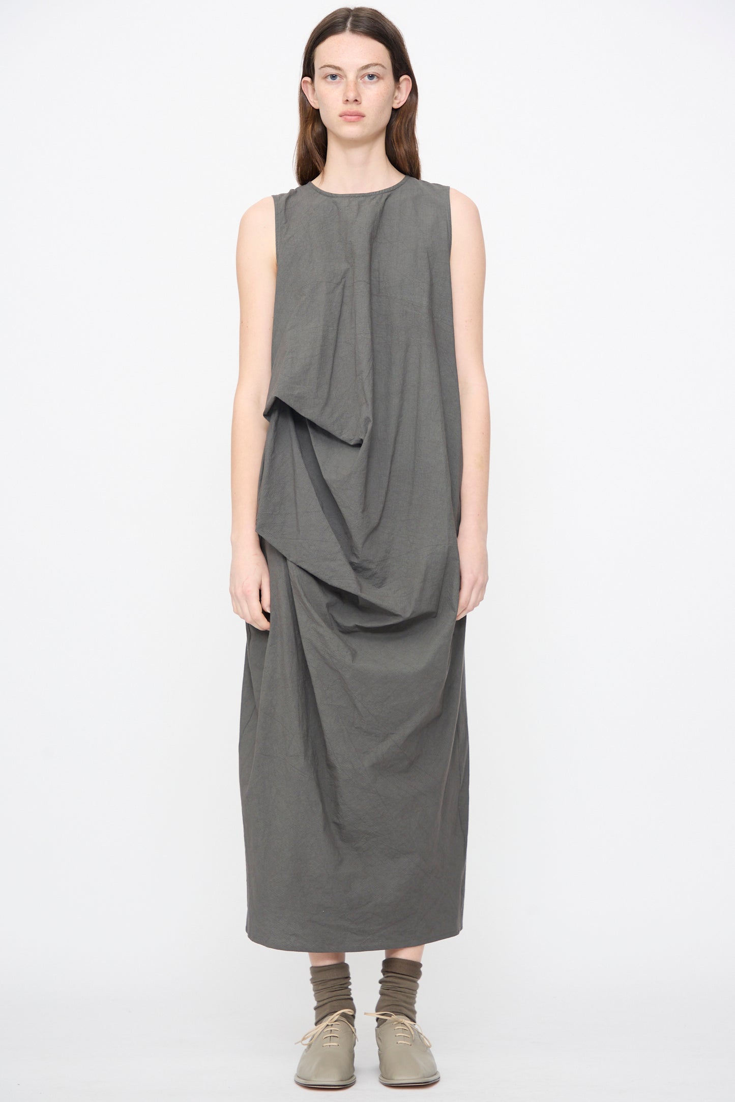 A person stands against a plain background wearing the Lauren Manoogian Ripstop Dress in Coal, a sleeveless midi with an asymmetrical gathered silhouette, paired with beige shoes and brown socks.