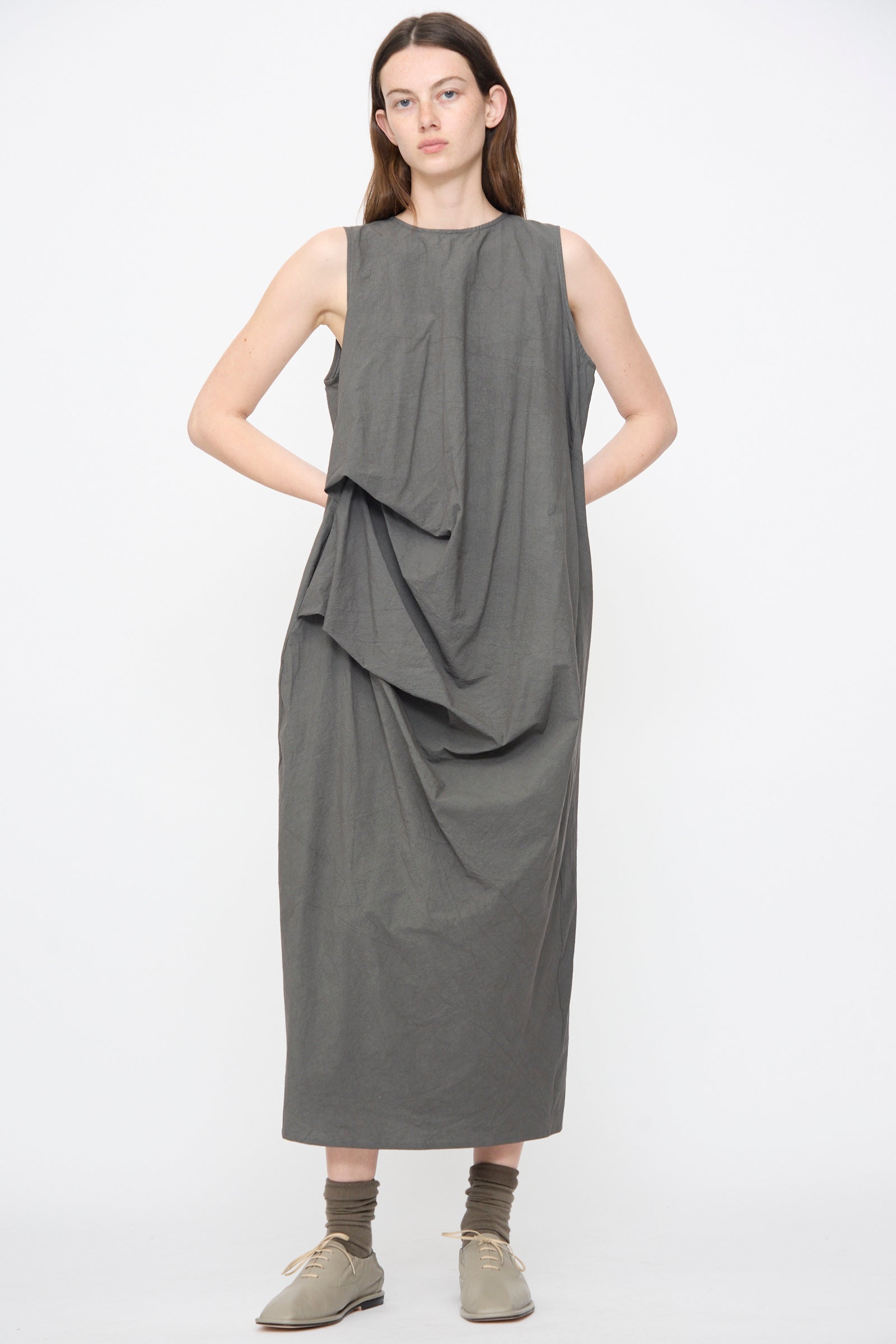 A woman faces forward wearing the Lauren Manoogian Ripstop Dress in Coal, a sleeveless dark grey cotton midi with an asymmetrical gathered silhouette, paired with tan lace-up shoes and olive socks.