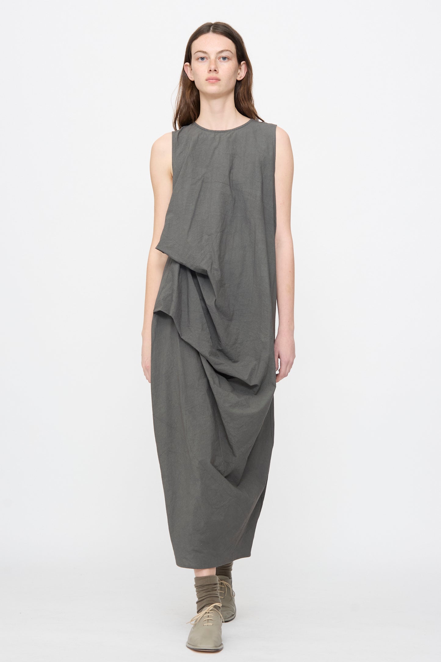 A person wears the Lauren Manoogian Ripstop Dress in Coal—a loose, sleeveless cotton dress with an asymmetrical gathered silhouette—paired with flat shoes and socks, standing against a plain white background.