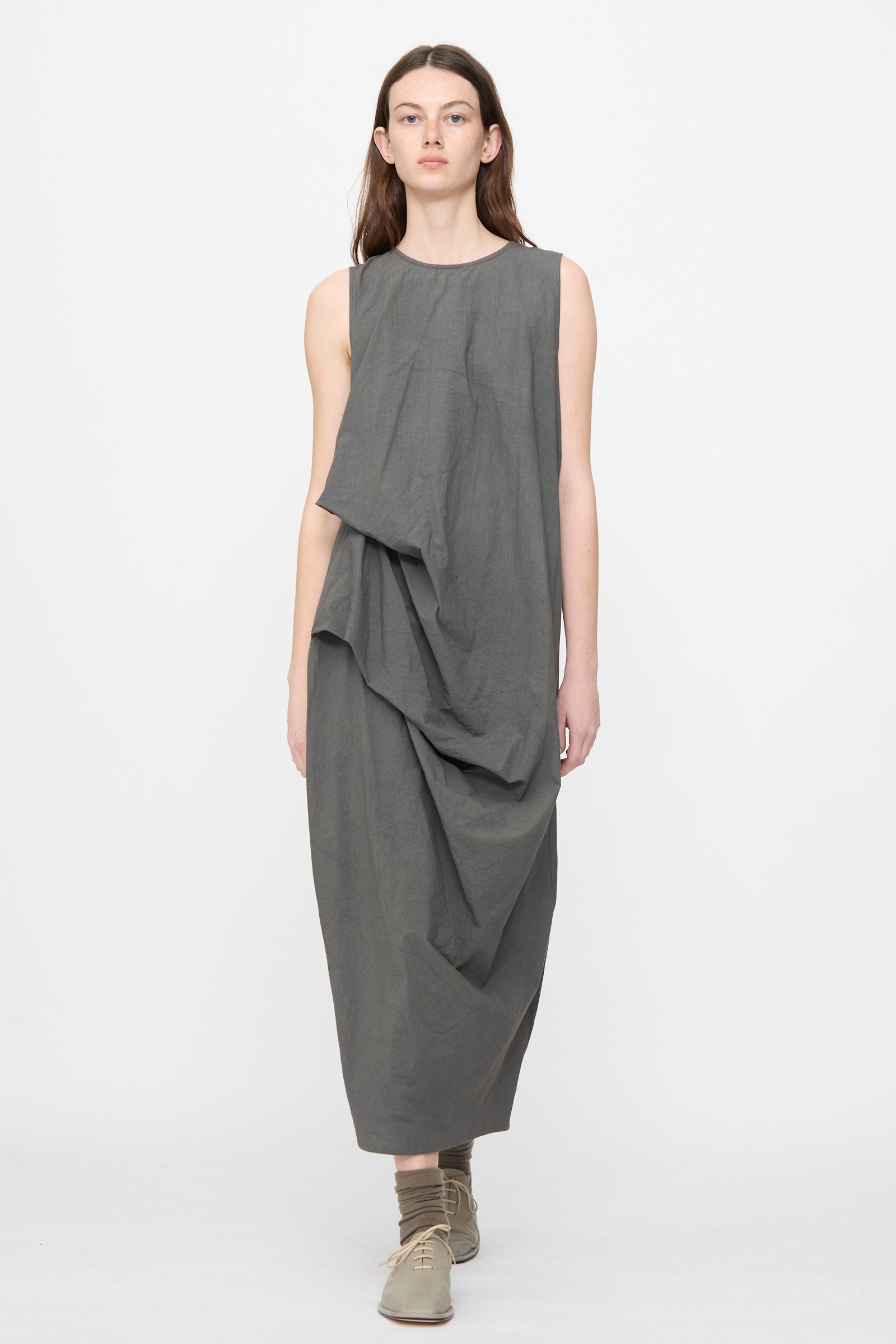 A person wears the Lauren Manoogian Ripstop Dress in Coal—a loose, sleeveless cotton dress with an asymmetrical gathered silhouette—paired with flat shoes and socks, standing against a plain white background.