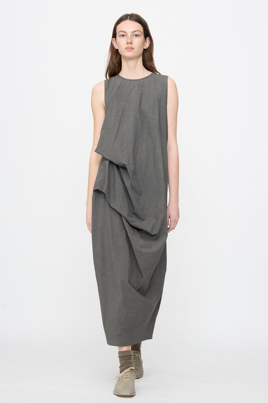 A person wears the Lauren Manoogian Ripstop Dress in Coal—a loose, sleeveless cotton dress with an asymmetrical gathered silhouette—paired with flat shoes and socks, standing against a plain white background.