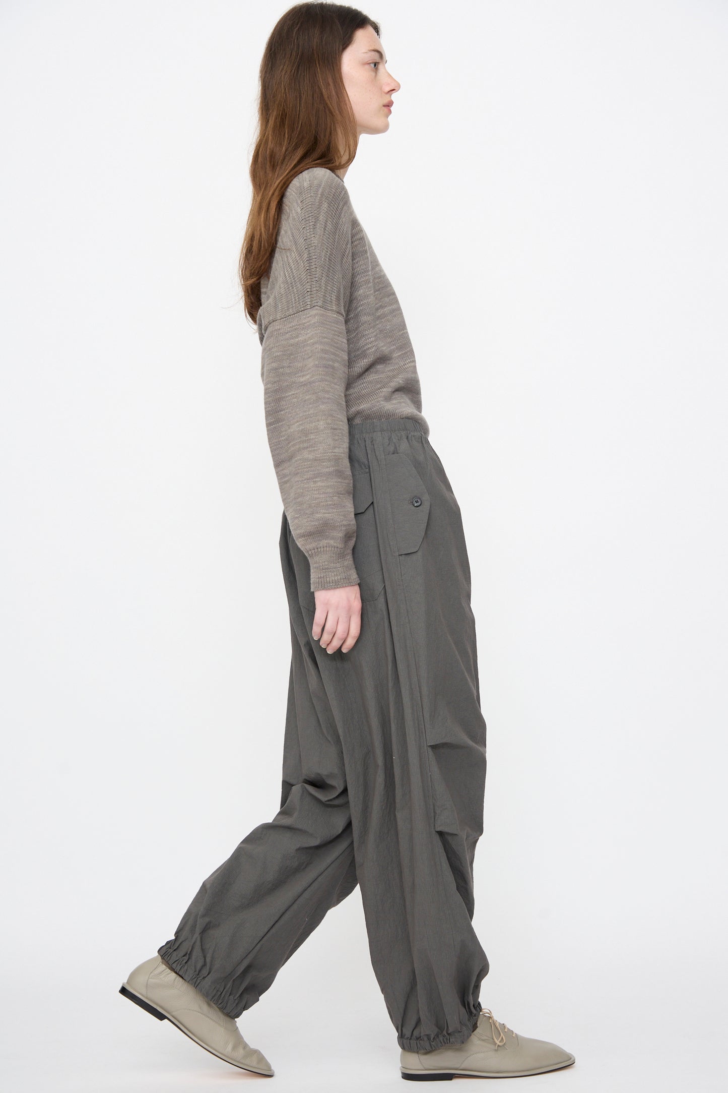 A person with long brown hair, wearing a gray sweater, Lauren Manoogian Ripstop Pants in Coal, and light gray shoes, walks against a plain white background.