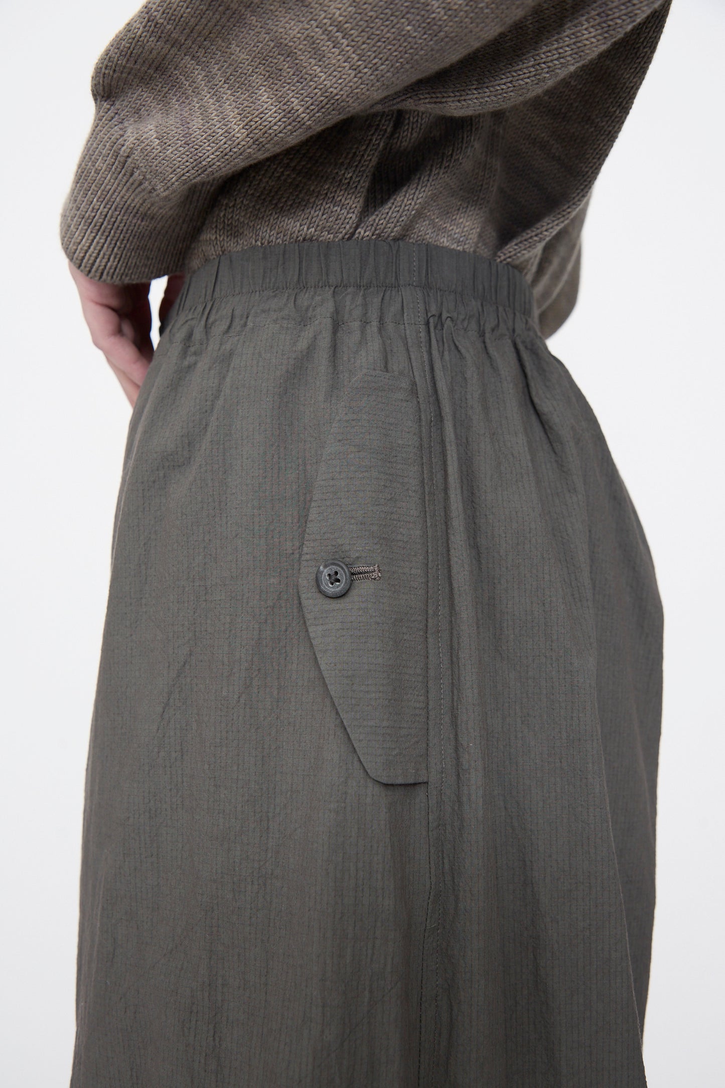 Close-up of a person's lower torso wearing a textured gray sweater and Lauren Manoogian Ripstop Pants in Coal, featuring an elastic waistband and buttoned pocket.