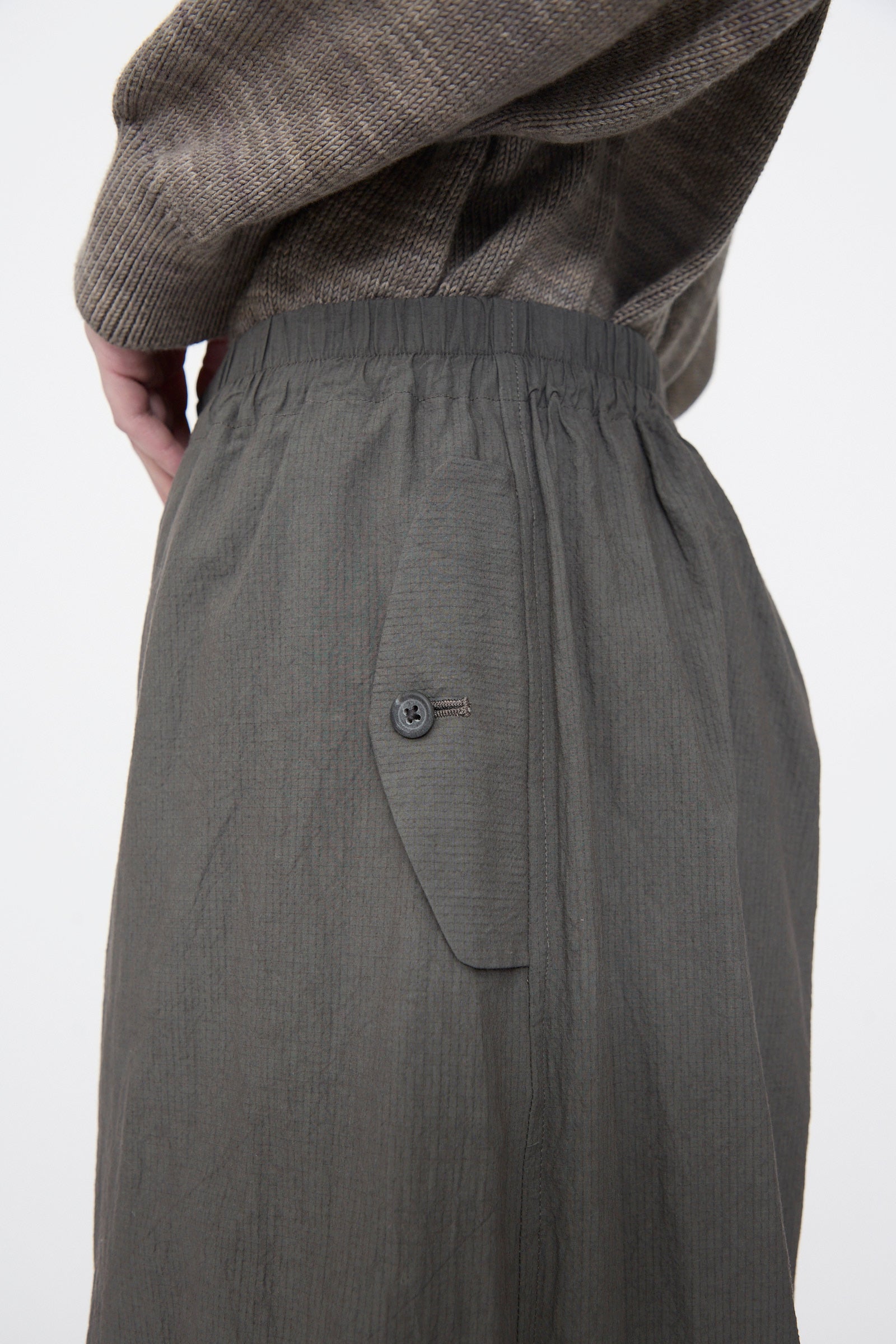 Close-up of a person's lower torso wearing a textured gray sweater and Lauren Manoogian Ripstop Pants in Coal, featuring an elastic waistband and buttoned pocket.