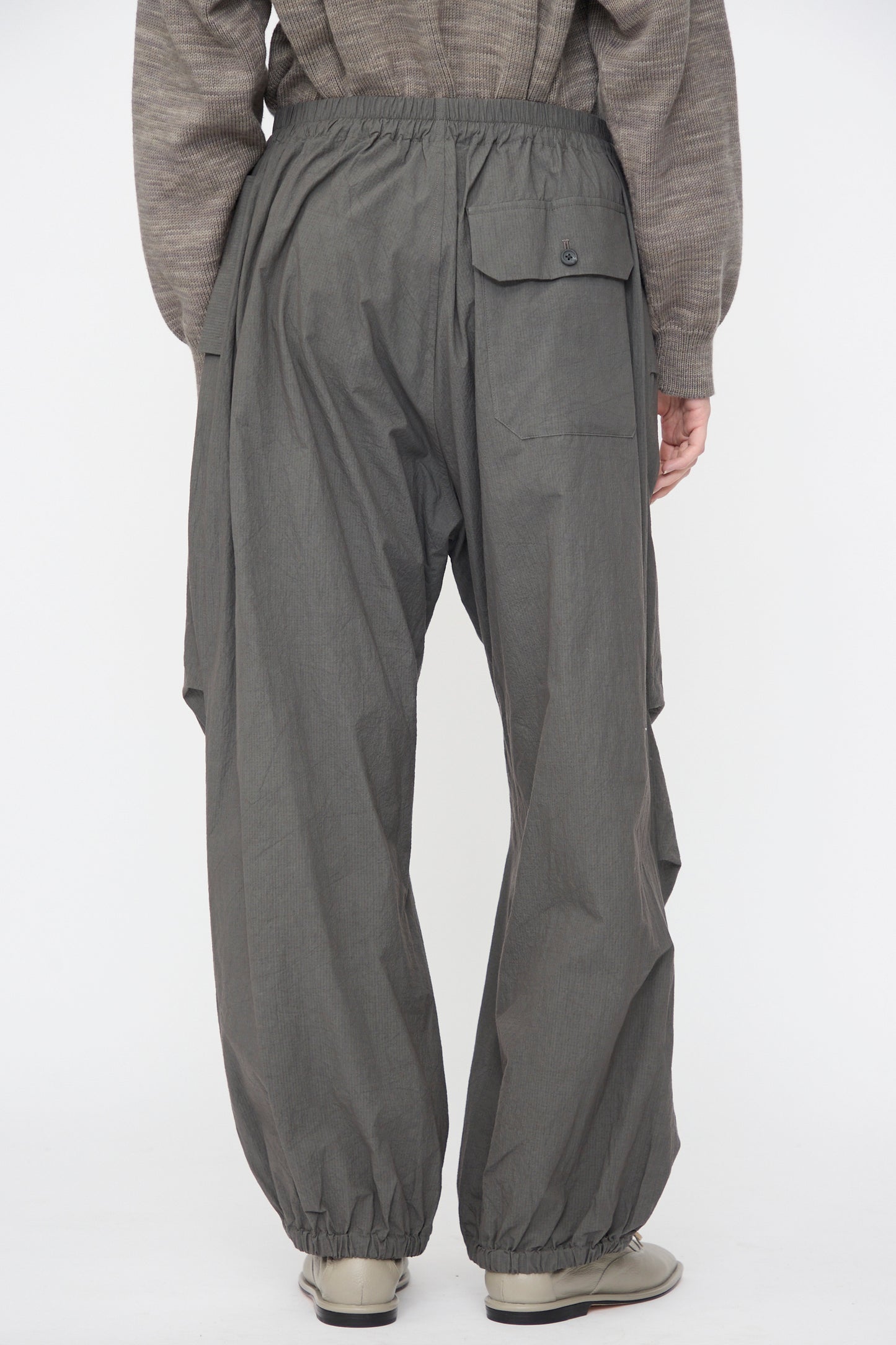 A person is seen from the back wearing Lauren Manoogian Ripstop Pants in Coal with an elastic waistband and ankles, paired with a gray textured top and light-colored shoes.
