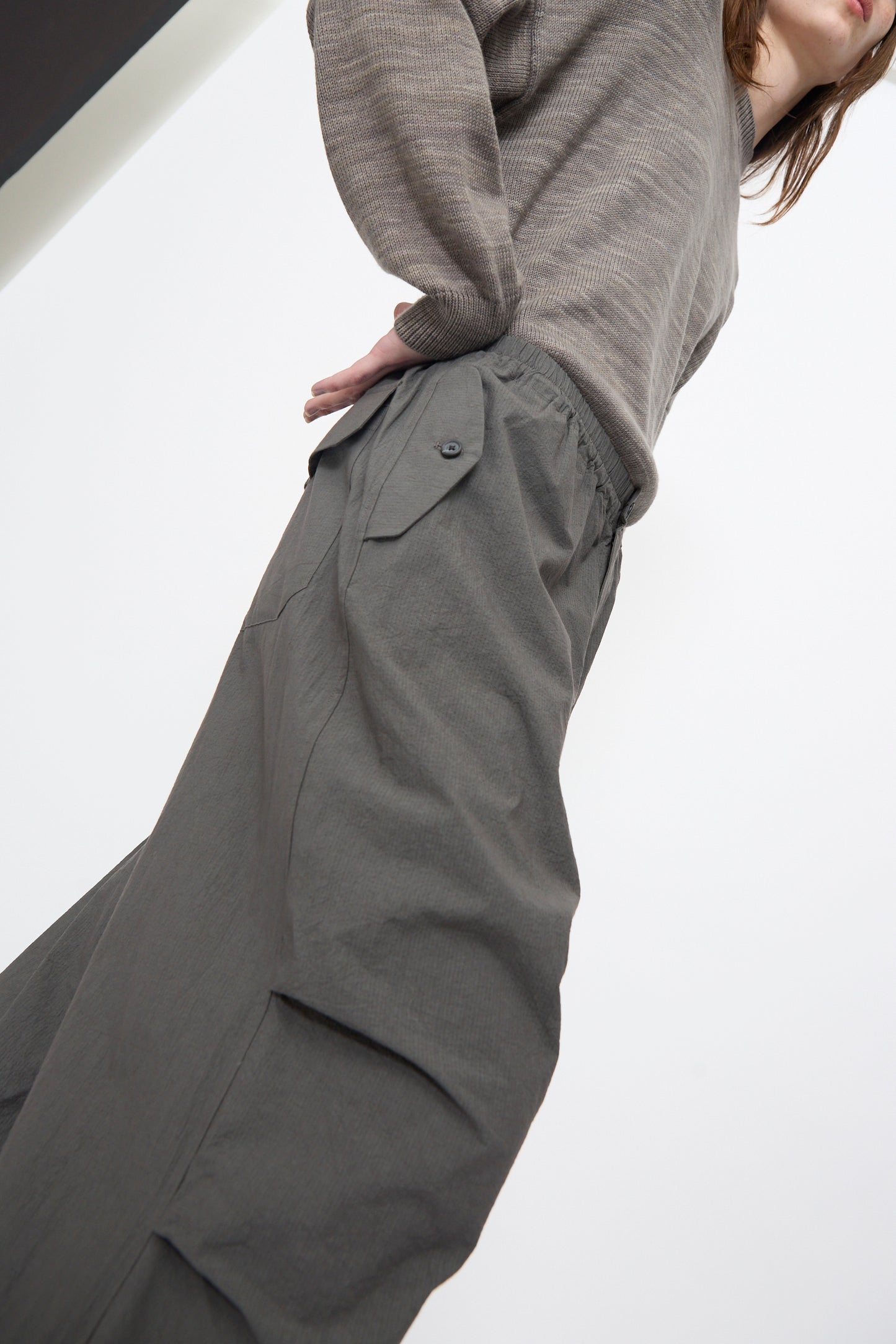 A person wears Lauren Manoogian's Ripstop Pants in Coal—loose, grey, cotton with an elastic waistband and side pocket—paired with a taupe long-sleeve top, photographed from a low angle against a white background.