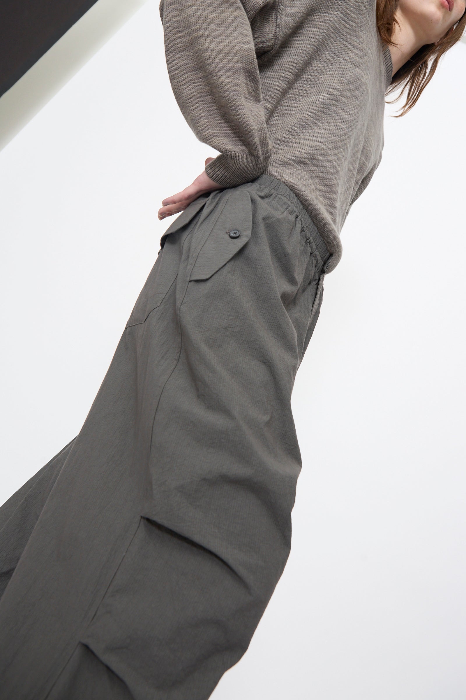 A person wears Lauren Manoogian's Ripstop Pants in Coal—loose, grey, cotton with an elastic waistband and side pocket—paired with a taupe long-sleeve top, photographed from a low angle against a white background.