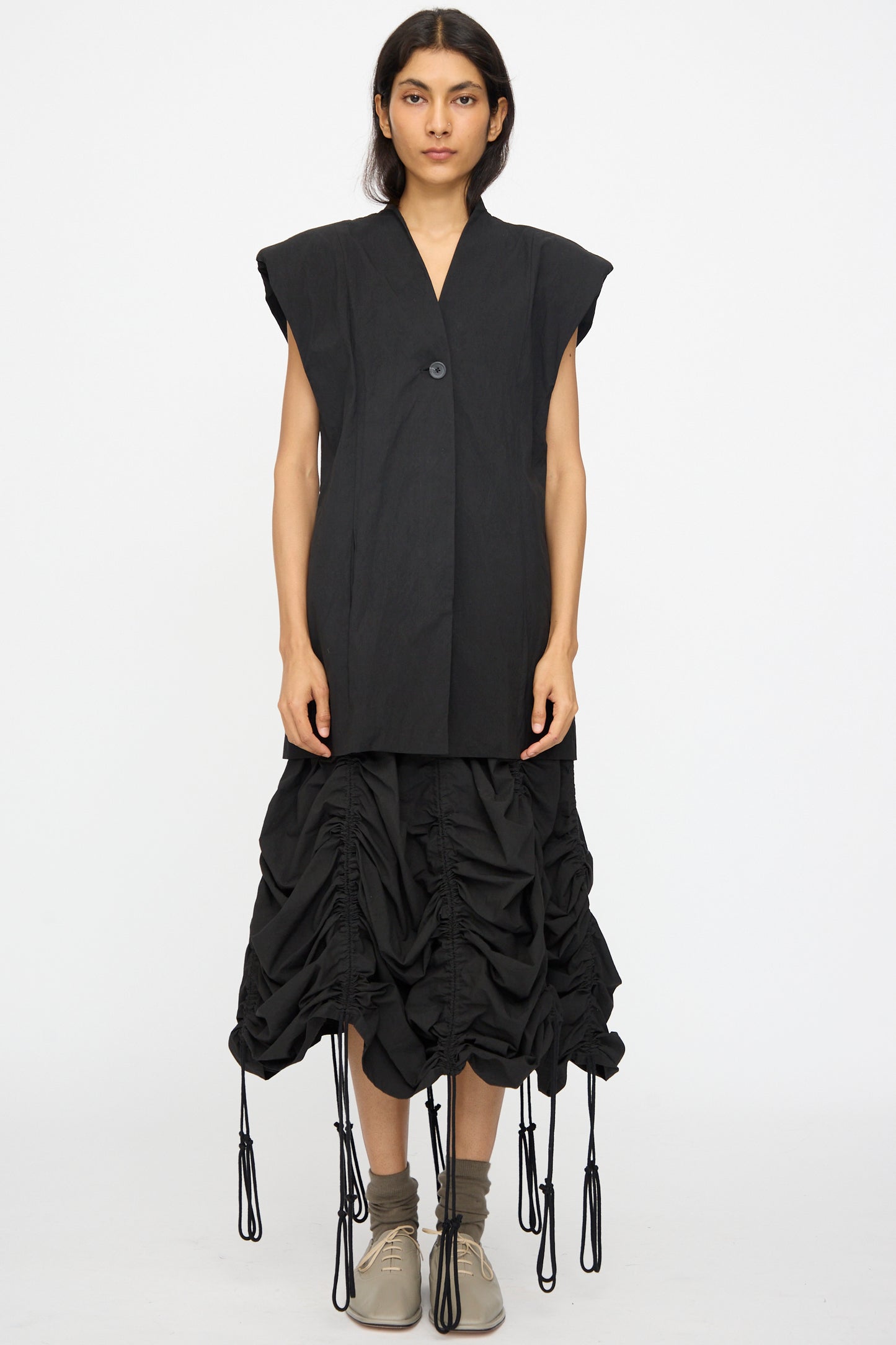A person faces forward against a plain white background wearing the Lauren Manoogian Shoulder Gilet in Black over a sleeveless black top, a ruched cotton-blend skirt with drawstrings, gray socks, and beige shoes.