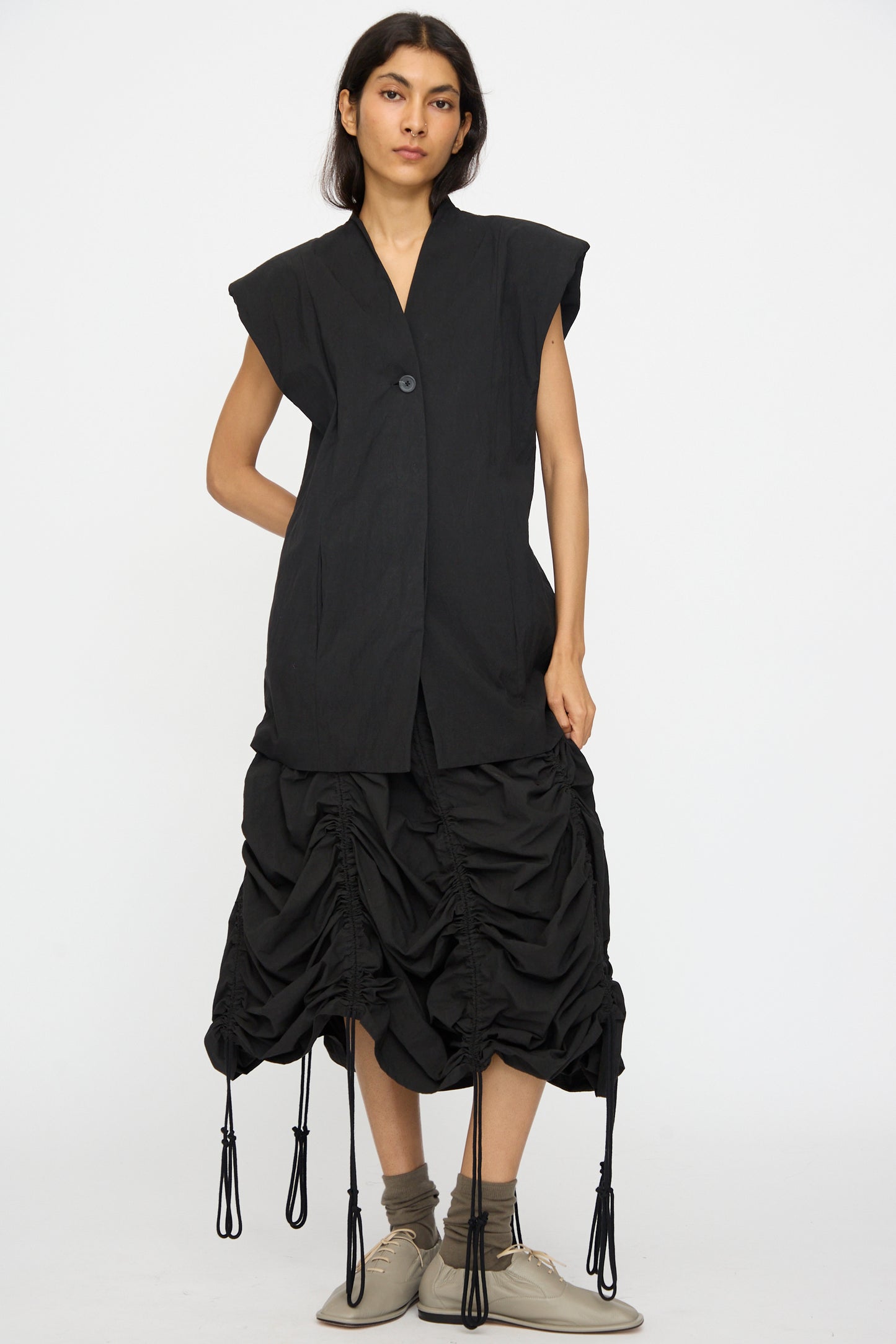 A person stands against a plain background wearing the Lauren Manoogian Shoulder Gilet in Black, paired with a black ruched skirt with drawstrings, gray socks, and beige shoes.