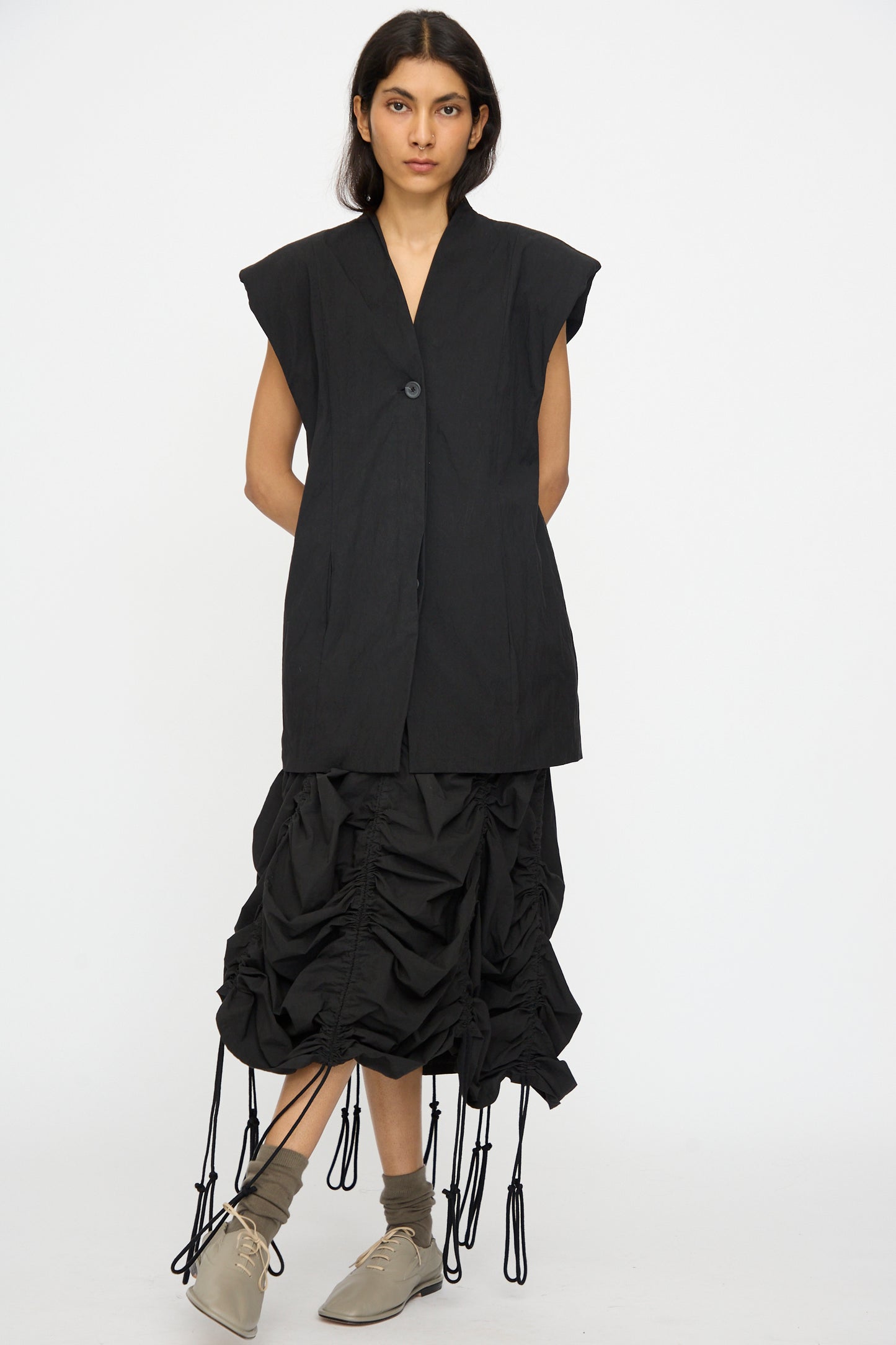 A woman stands against a plain background wearing the Lauren Manoogian Shoulder Gilet in Black, a black ruched skirt with hanging ties, beige socks, and light beige shoes.