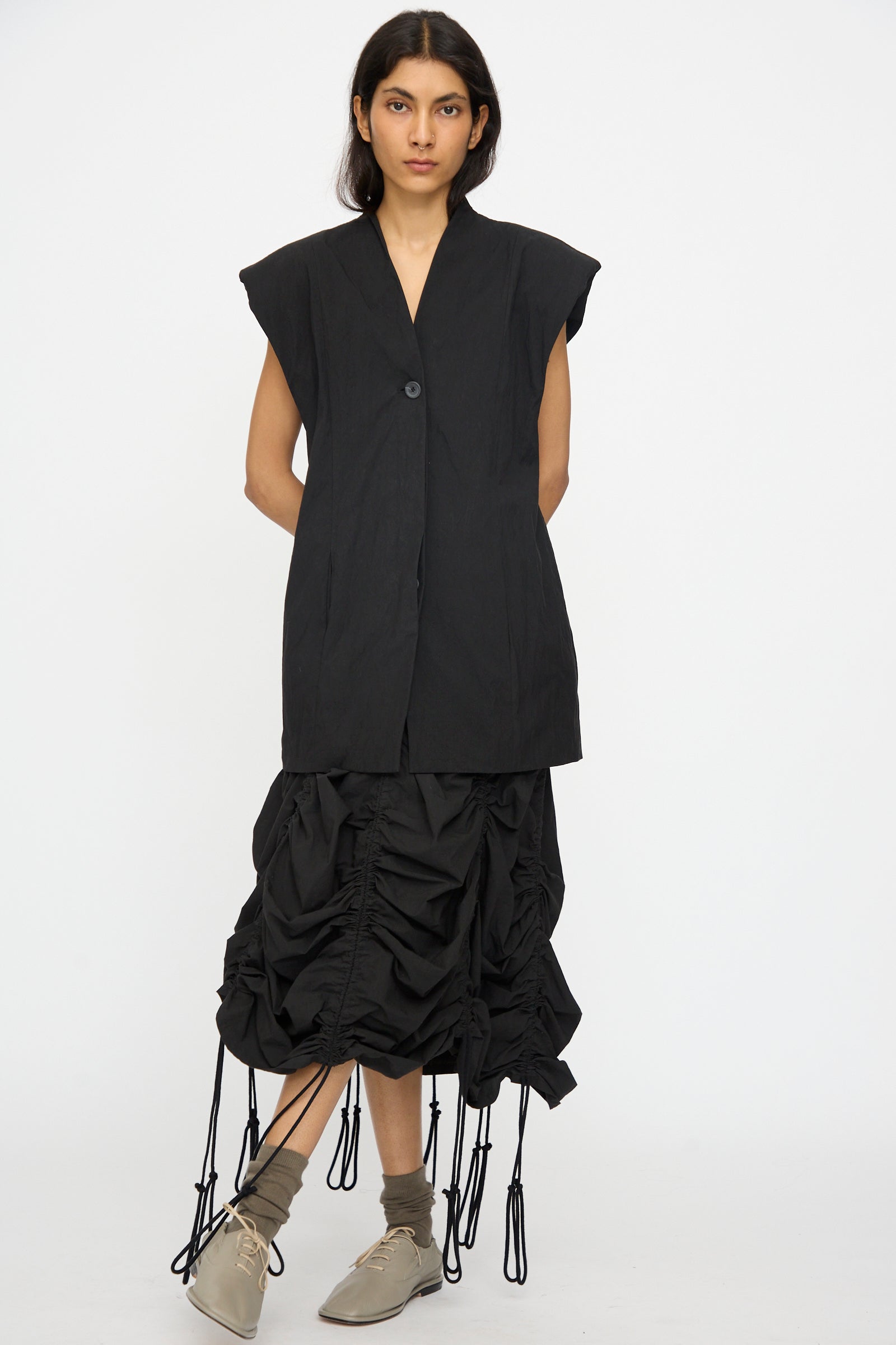 A woman stands against a plain background wearing the Lauren Manoogian Shoulder Gilet in Black, a black ruched skirt with hanging ties, beige socks, and light beige shoes.