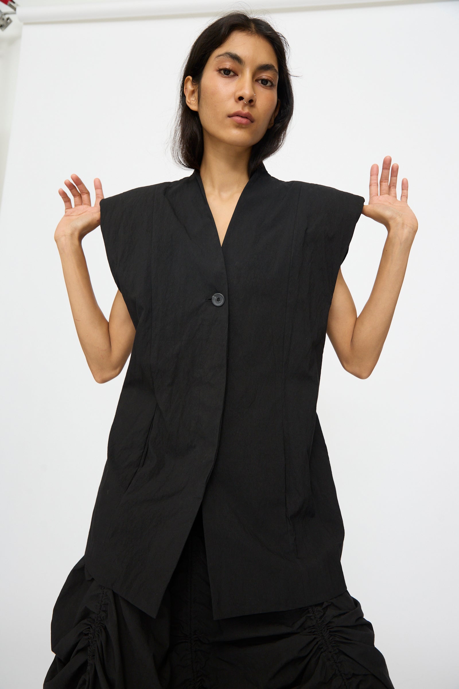 A woman poses against a white background wearing the Lauren Manoogian Shoulder Gilet in Black, a sleeveless, oversized cotton-blend piece with a single button, holding her hands up near her shoulders.