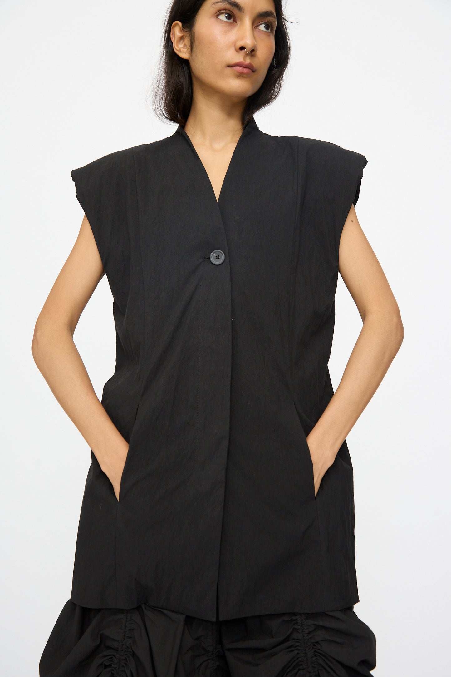 A woman models the Lauren Manoogian Shoulder Gilet in Black—a sleeveless, single-button cotton blend—standing hands in pockets against a plain white background.