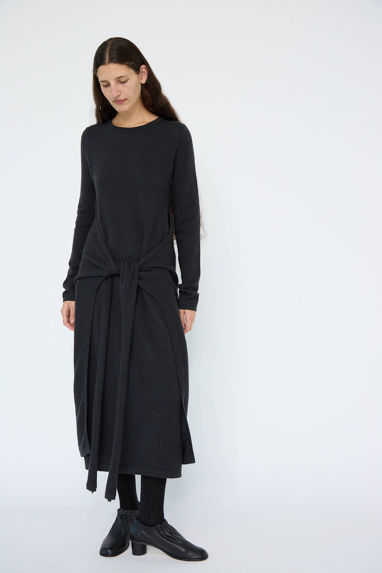 A person in the Lauren Manoogian Sleeves Dress in Ink, featuring a long-sleeve design made from a dark gray wool/alpaca blend with a front knot detail, stands against a plain white background, paired with black ankle boots.