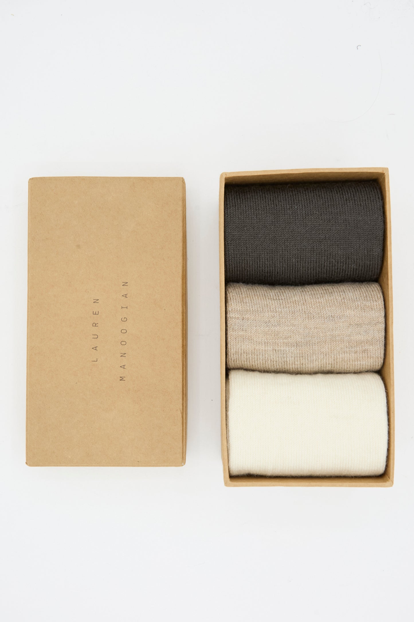 A partially open box reveals the Lauren Manoogian Sock Set in Mix—three neatly rolled pairs of alpaca wool socks in dark gray, beige, and off-white, made in Peru.