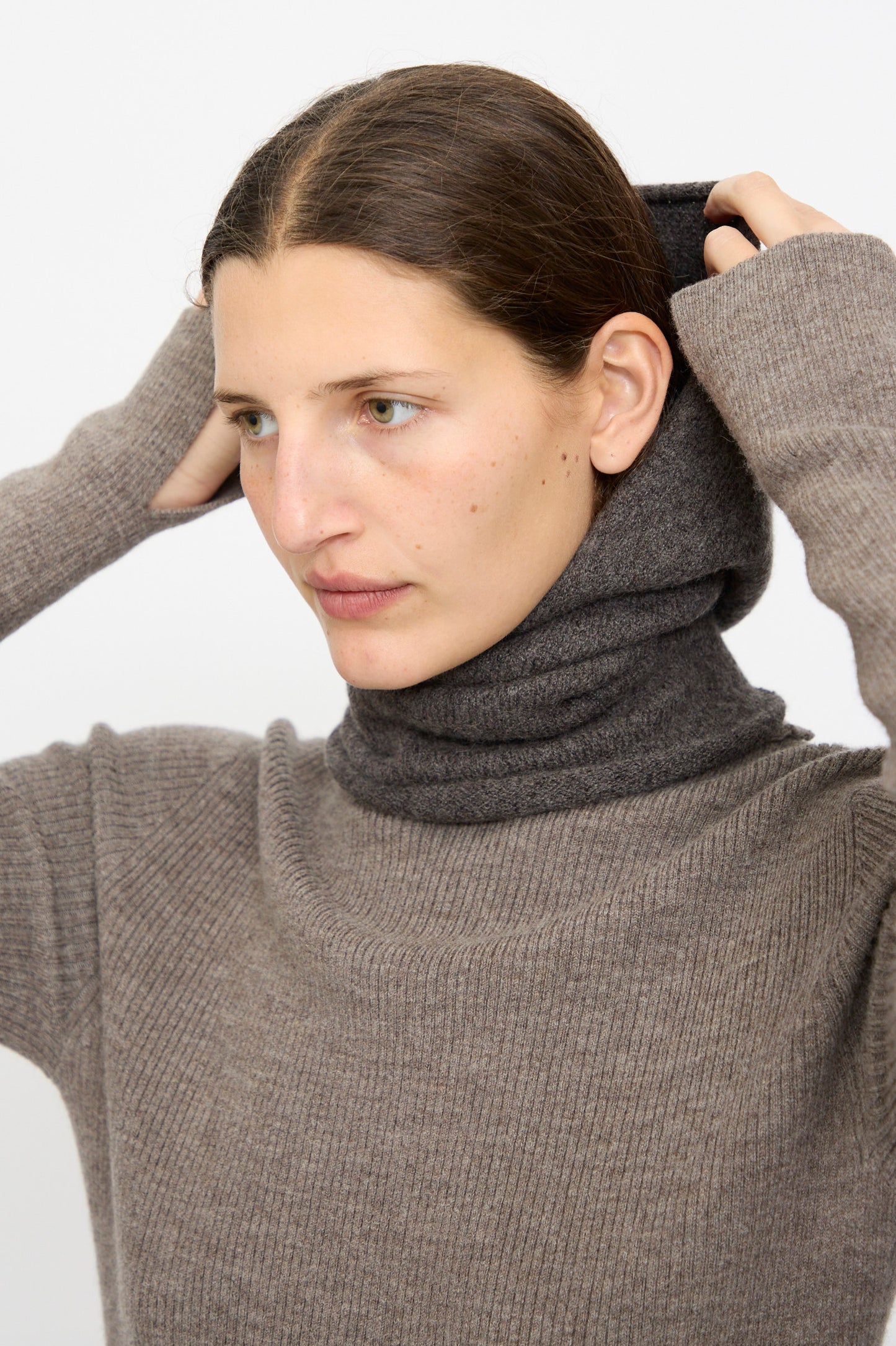 A person in a brown ribbed sweater pulls a Lauren Manoogian Soft Balaclava in Blackened over their head against a plain light background.