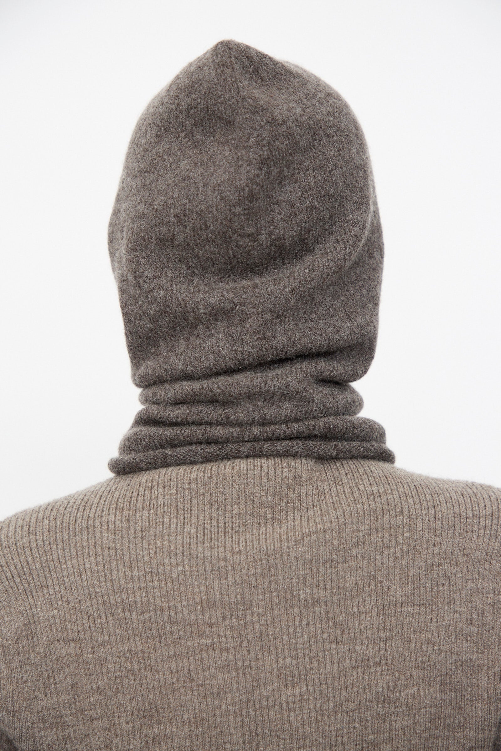 A person shown from the back against a plain white background wears a ribbed brown hooded sweater paired with the Lauren Manoogian Soft Balaclava in Blackened.