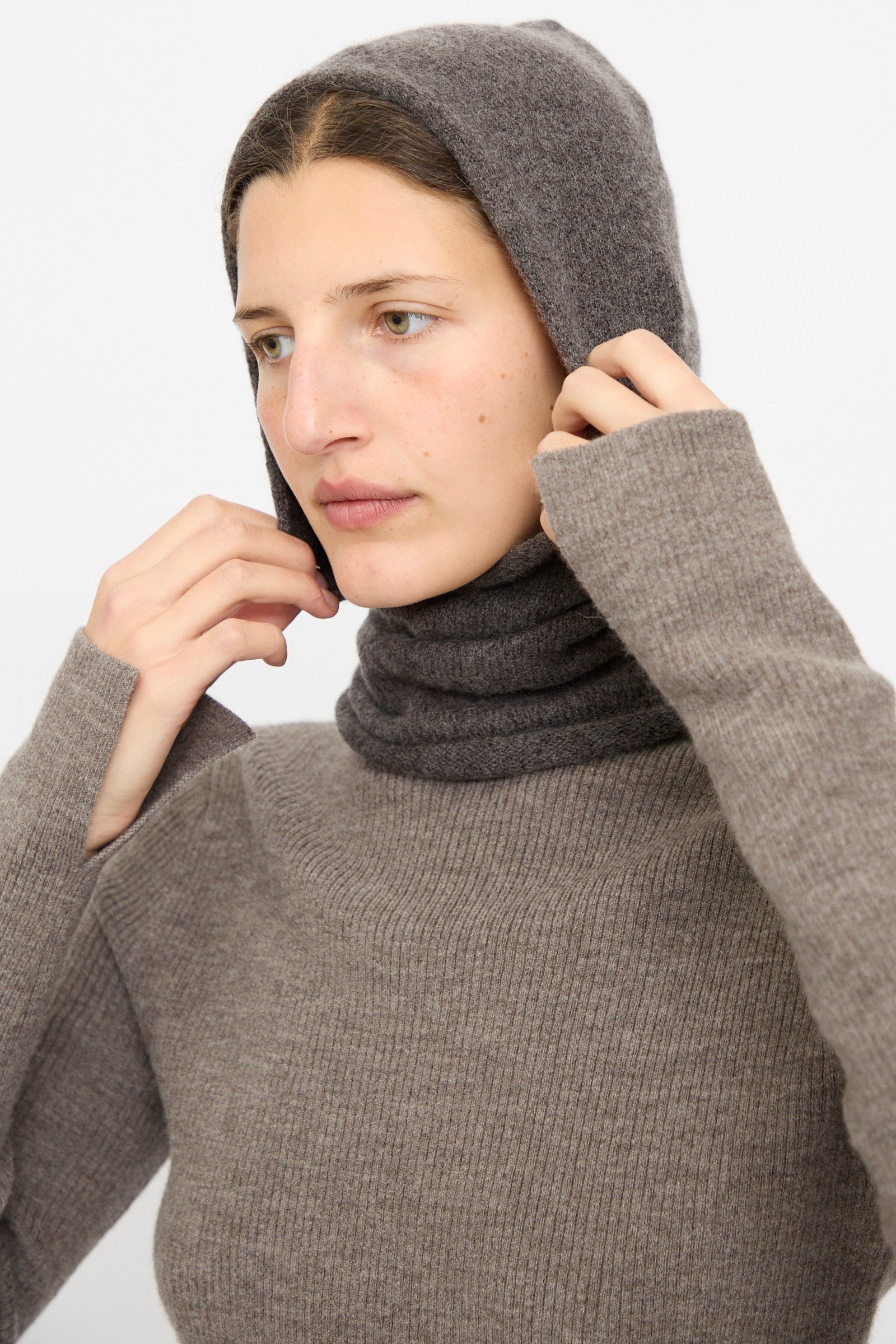 A person wearing the Lauren Manoogian Soft Balaclava in Blackened, adjusting the hood with both hands over their head.