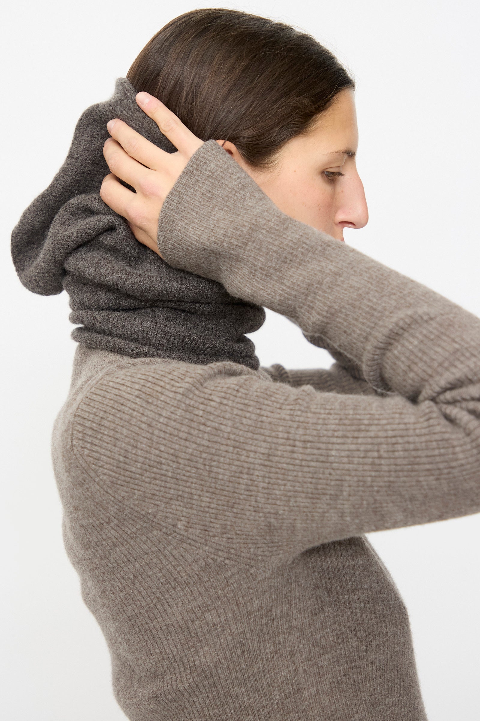 A person wearing the Lauren Manoogian Soft Balaclava in Blackened adjusts the collar over their head, shown in profile against a plain light backdrop—capturing the cozy feel of this ribbed knit accessory.