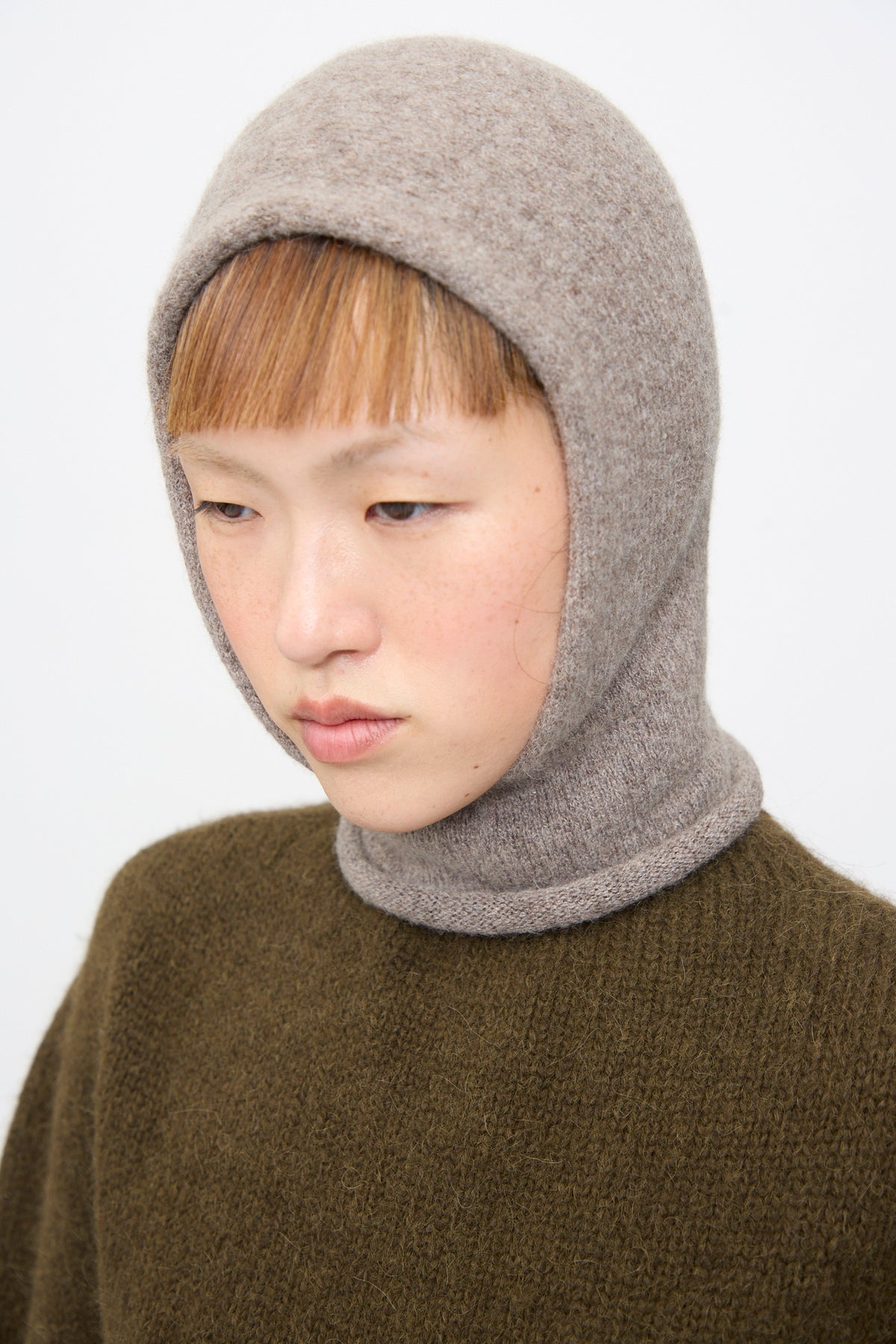 A person wearing a handloomed Lauren Manoogian Soft Balaclava in Grey Melange over a dark olive-green sweater.