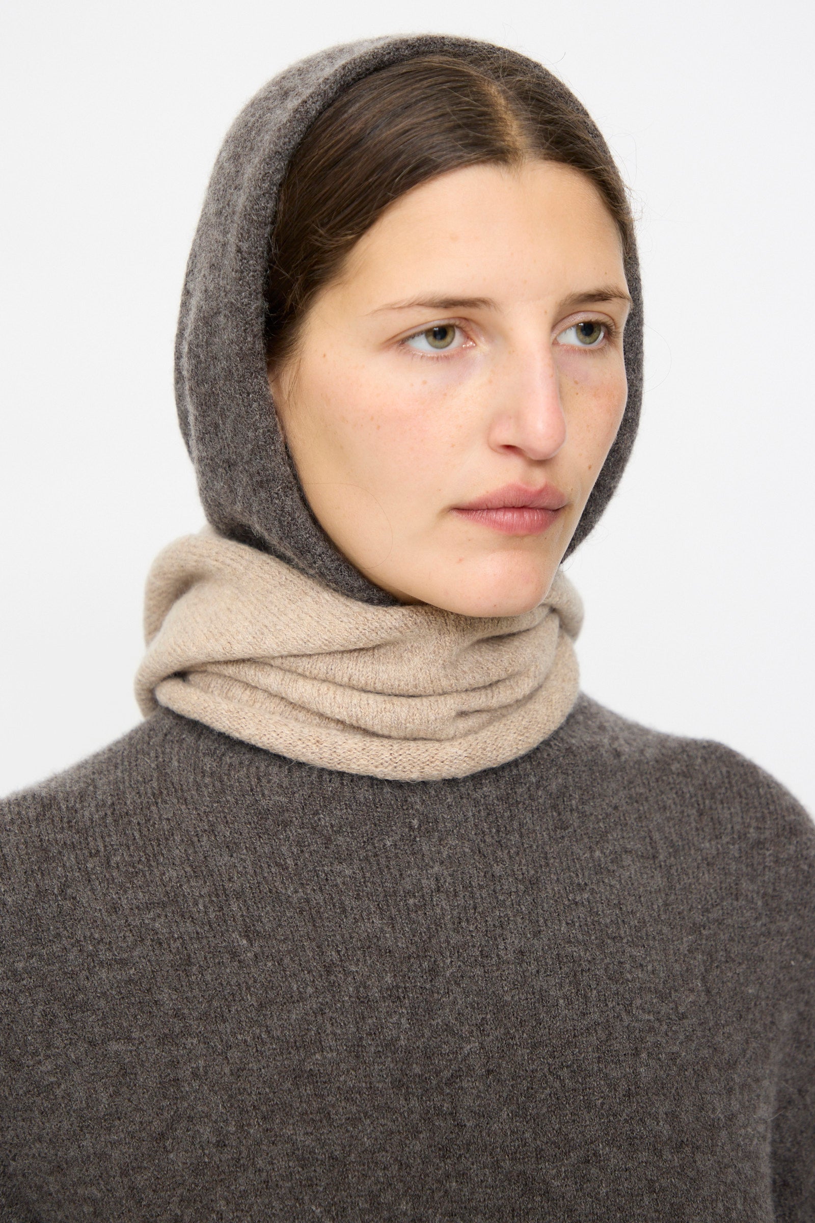 A person with light skin and brown hair wears a brown knit hooded sweater and the Lauren Manoogian Soft Balaclava in Oak, made in Peru, wrapped around their neck against a plain background.