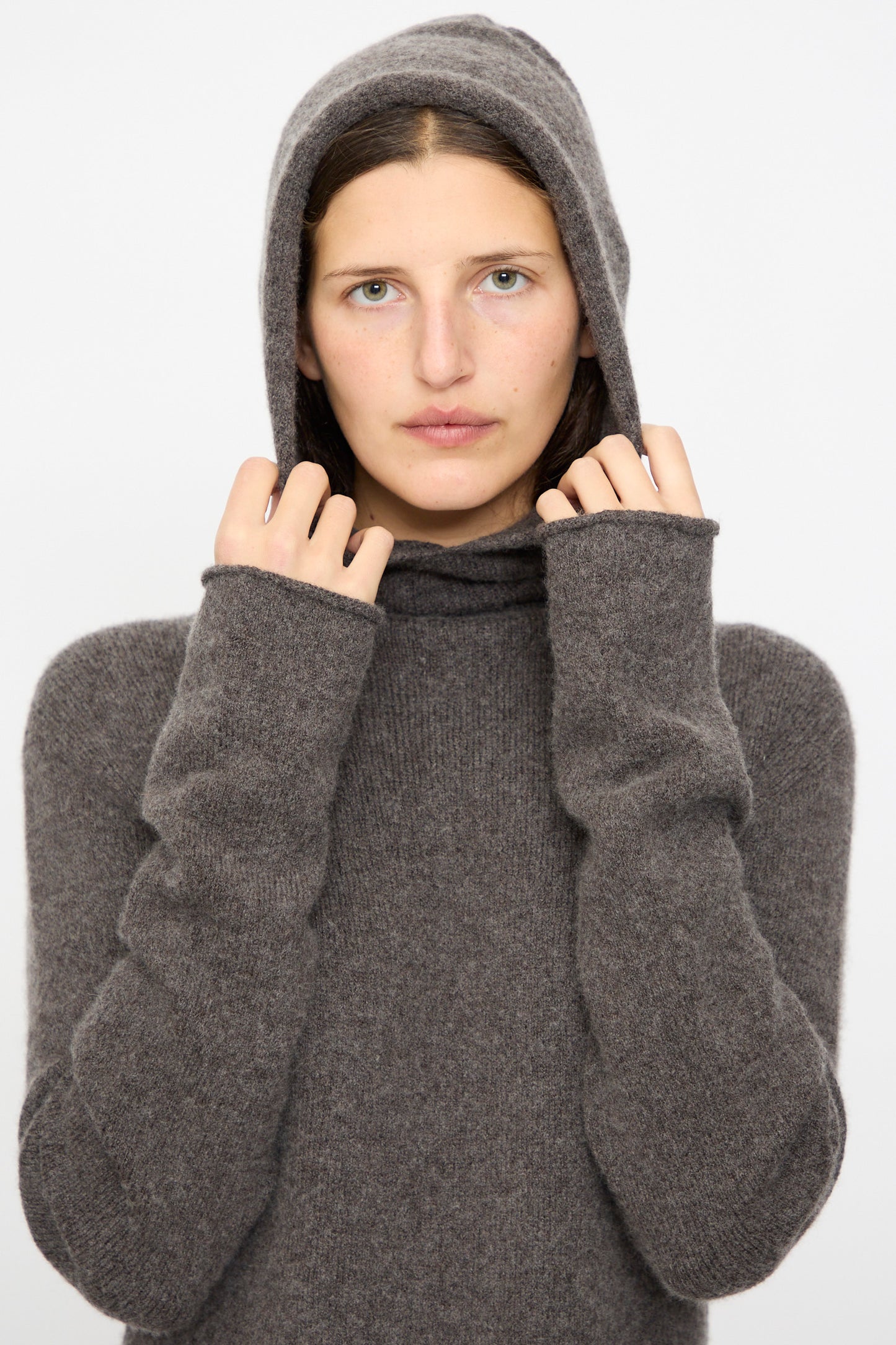 Model wears the Lauren Manoogian Soft Balaclava Dress in Blackened, looking at the camera and holding the hood, set against a plain background.