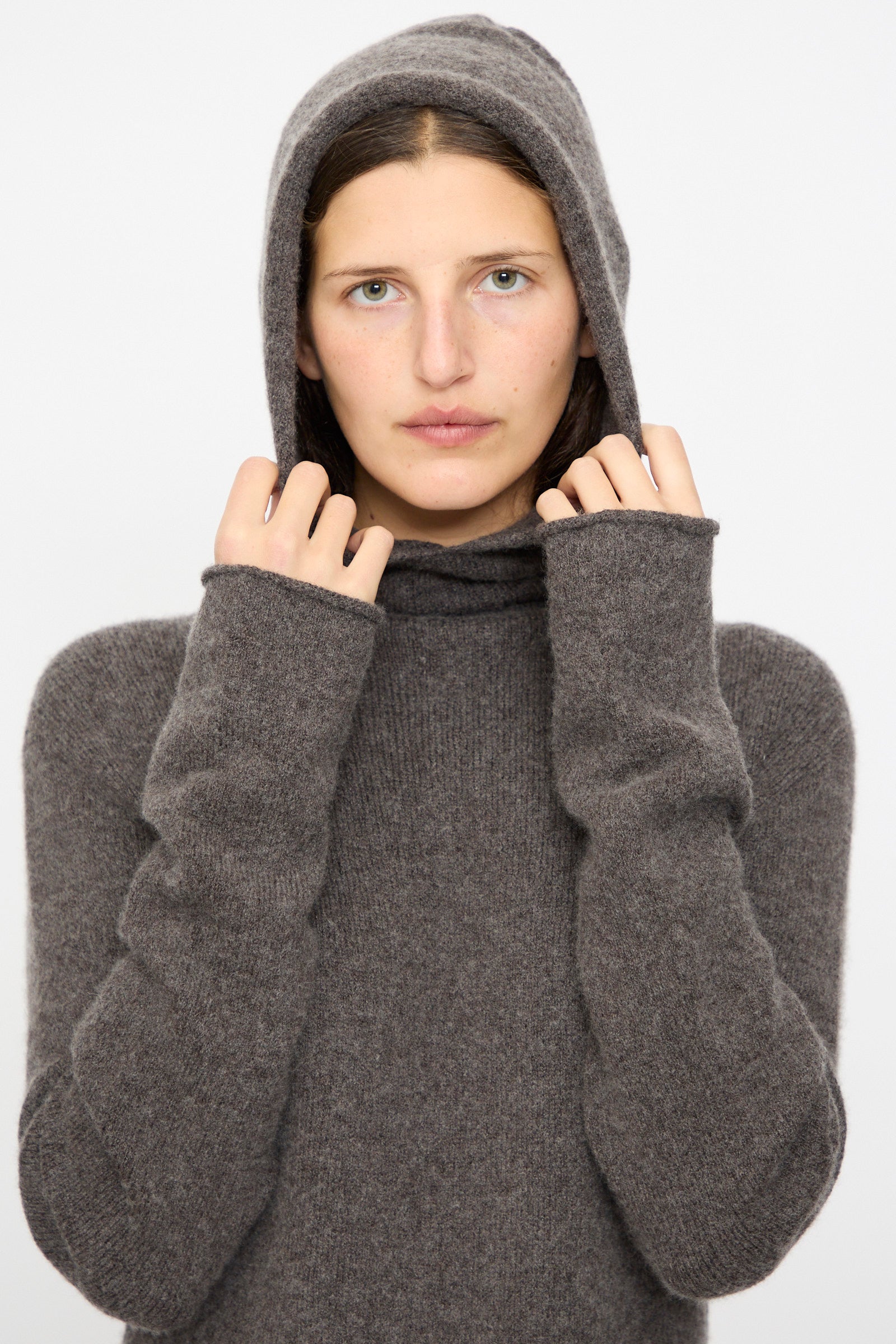 Model wears the Lauren Manoogian Soft Balaclava Dress in Blackened, looking at the camera and holding the hood, set against a plain background.