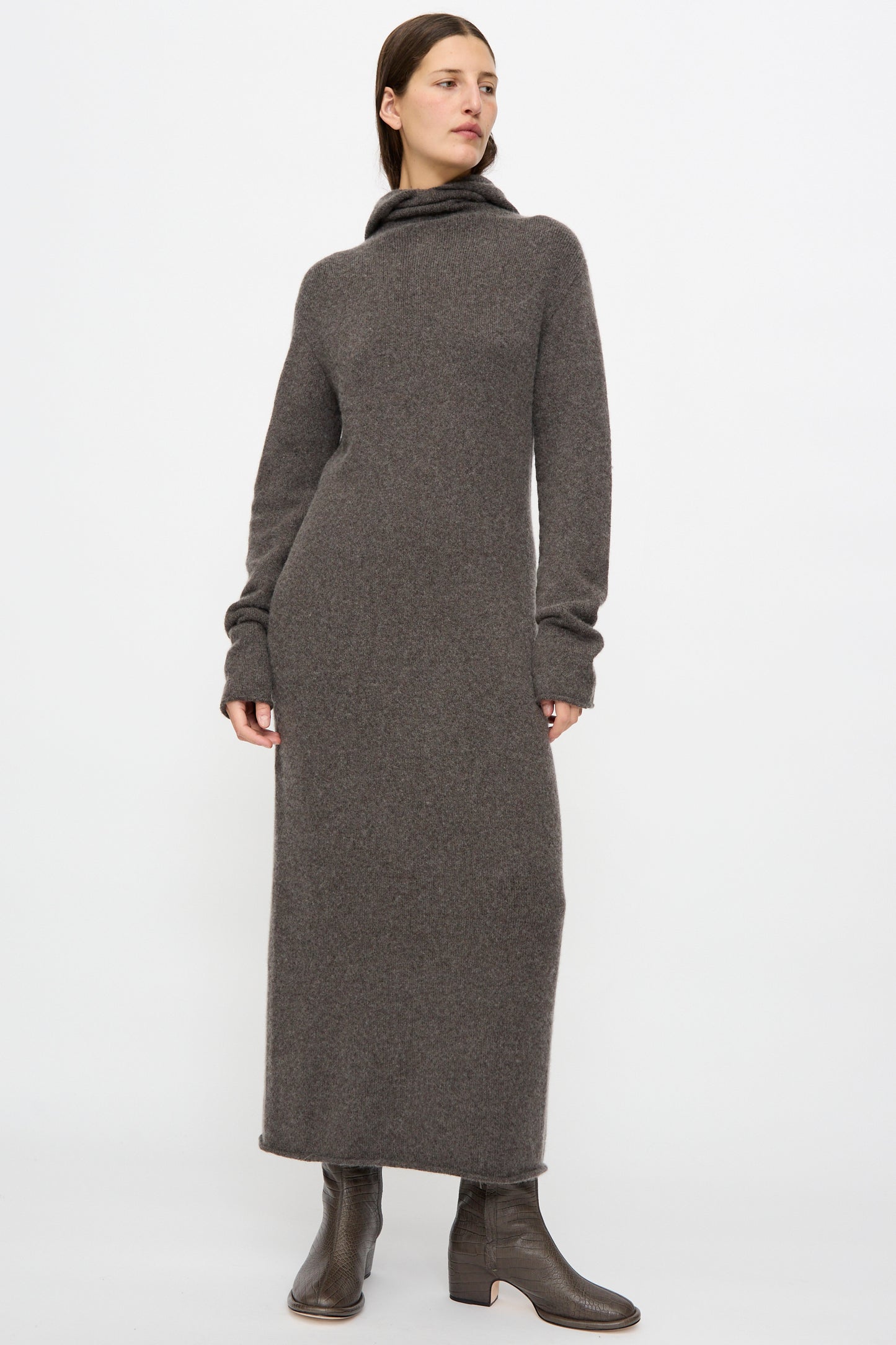 A person stands against a plain white background wearing Lauren Manoogian's Soft Balaclava Dress in Blackened, a long baby alpaca maxi dress with a turtleneck and long sleeves, paired with brown ankle boots.