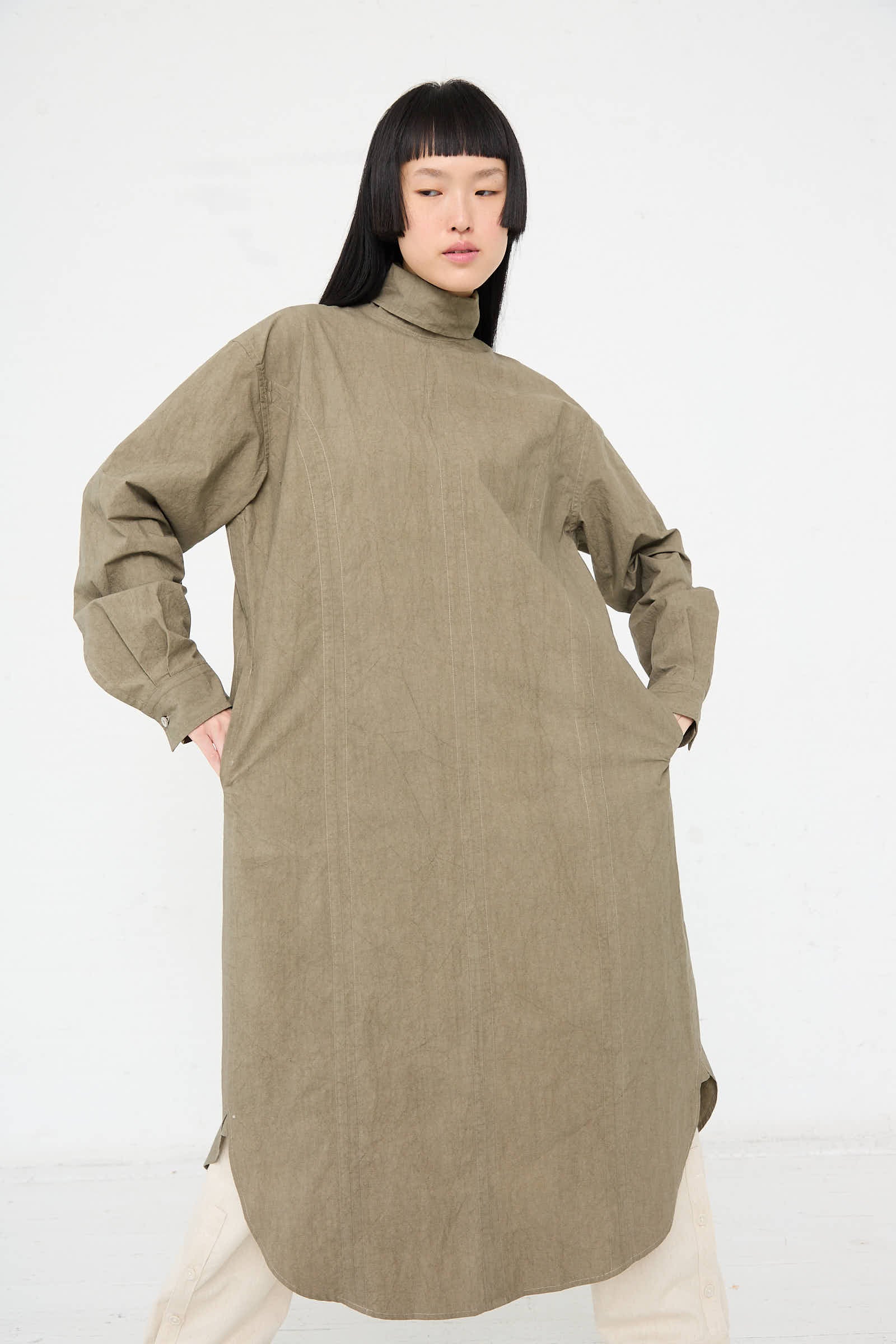 A person with long, dark hair wears the Lauren Manoogian Soft Collar Shirt Dress in Clay, featuring buttoned cuffs and a high neckline, as they pose with hands on hips against a white background.