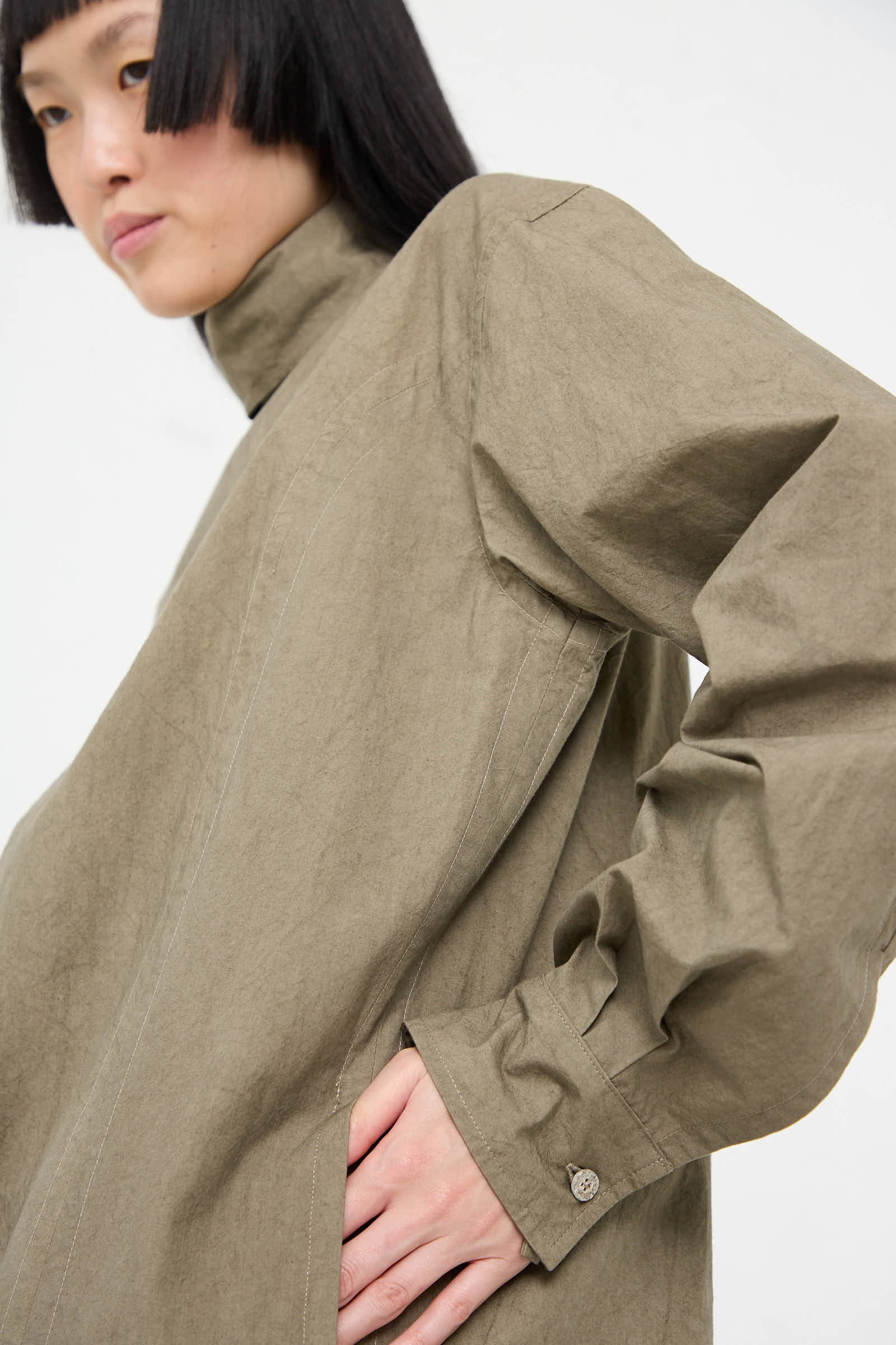 A person with short dark hair wears the Soft Collar Shirt Dress in Clay by Lauren Manoogian, a loose-fitting, high-collared, long-sleeved garment with buttoned cuffs, their hand resting on their hip.