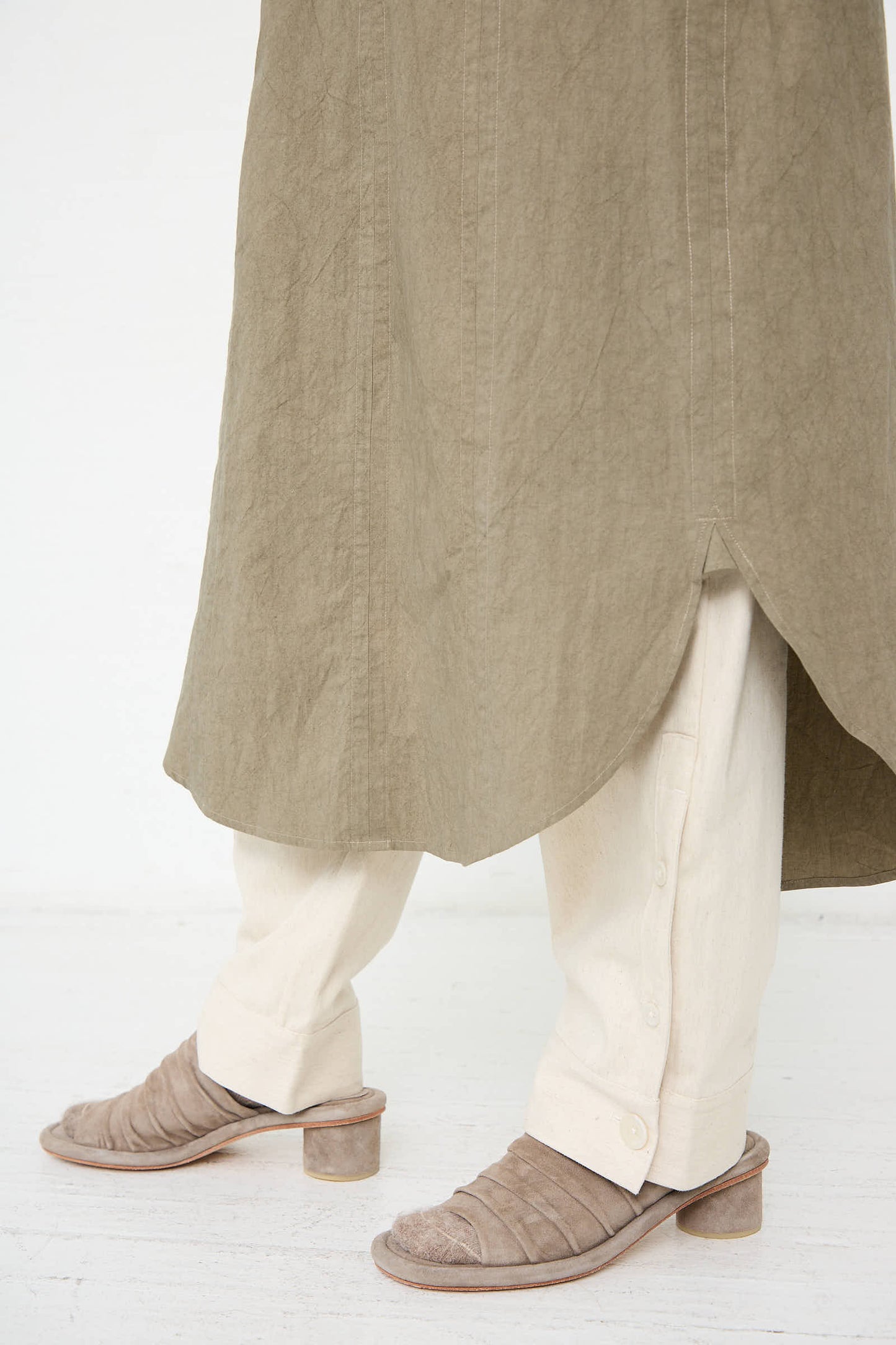 A person wearing a clay Soft Collar Shirt Dress by Lauren Manoogian over beige pants and brown heeled sandals, with buttoned cuffs, stands on a white floor.