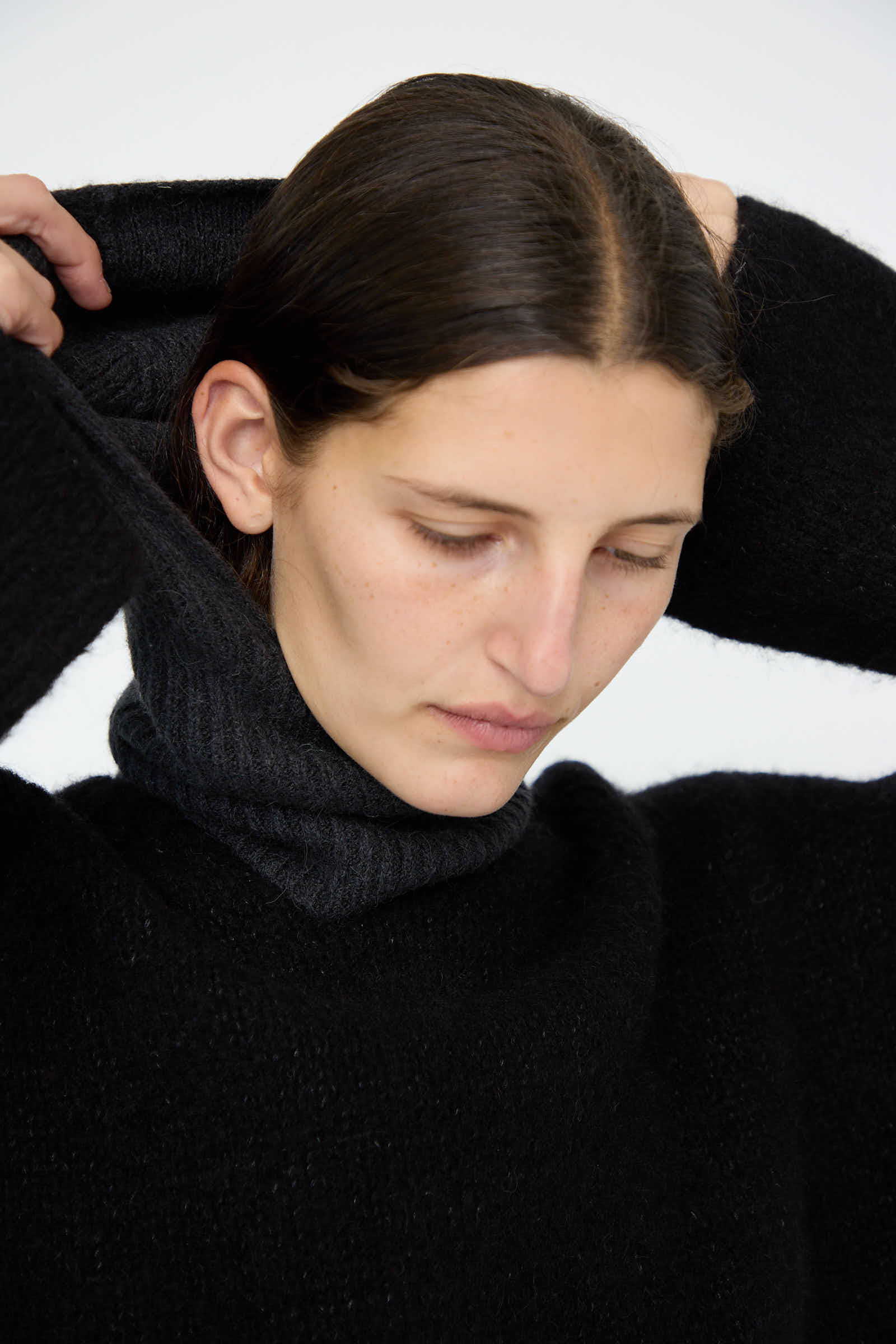 A person with dark hair adjusts a soft rib balaclava in ink by Lauren Manoogian against a plain background.