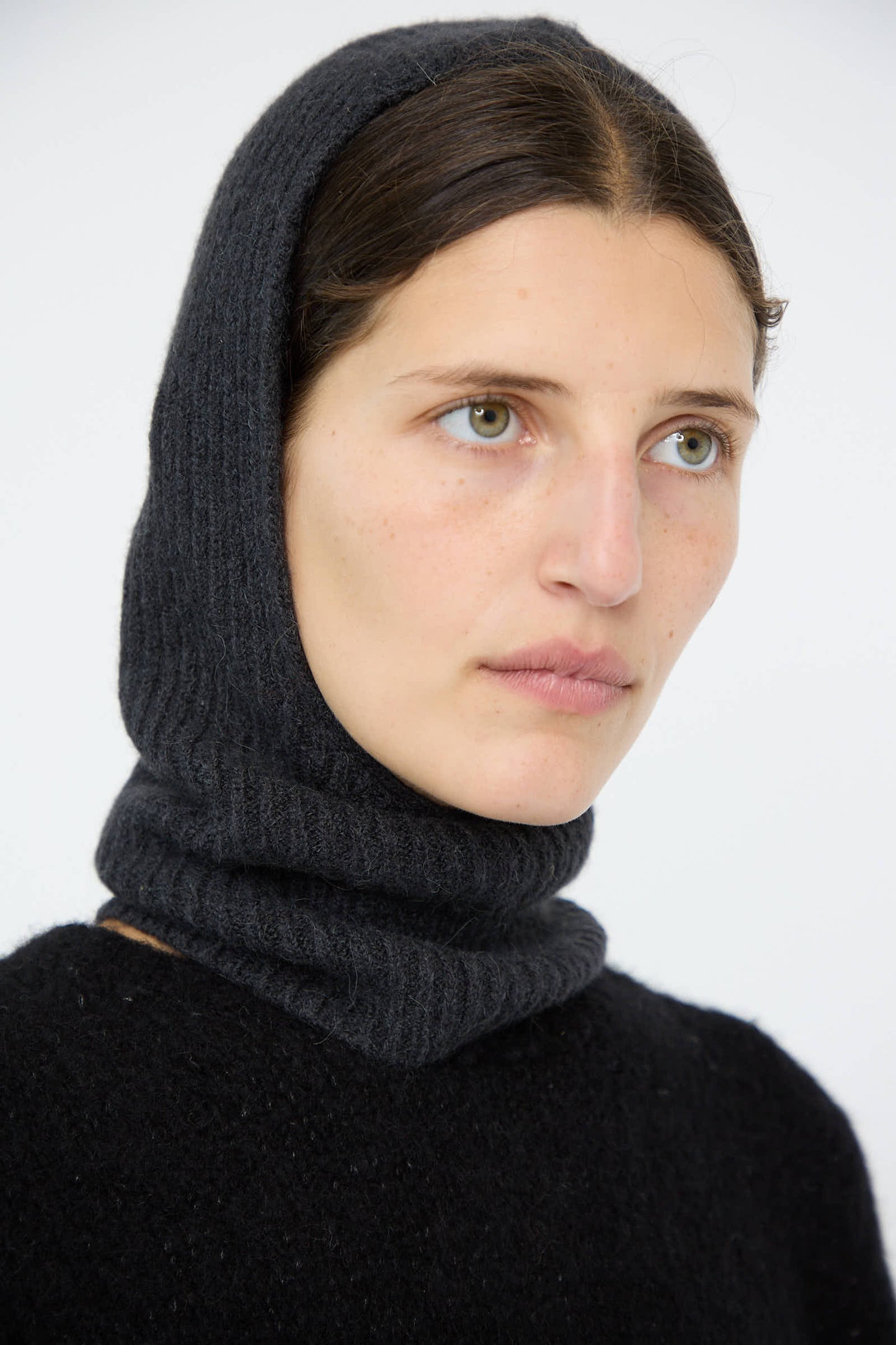 A person with long dark hair wears the Lauren Manoogian Soft Rib Balaclava in Ink and a matching sweater, looking to the side against a plain white background.