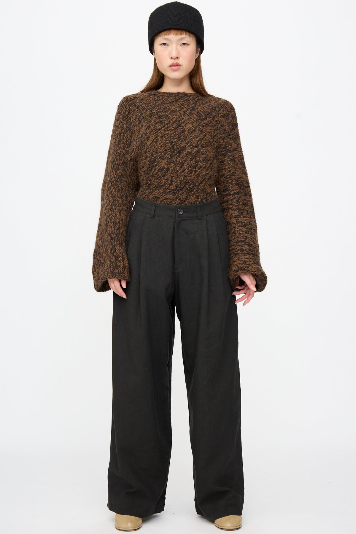 A person wearing a brown knit sweater, a black hat, and Lauren Manoogian Soft Trouser in Black pleated trousers.